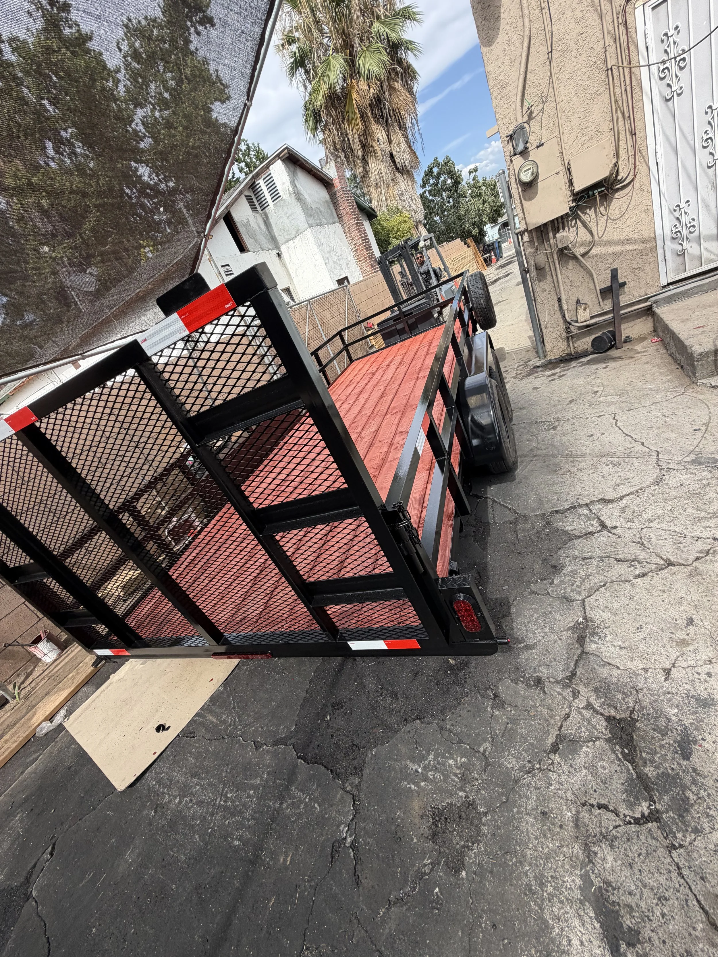 A flatbed utility trailer parked on a cracked asphalt driveway, with a black metal frame, a red wooden deck, and a black metal gate. In the background, a man is operating a forklift, and there is a beige building with electrical wires and an old palm tree with brown, drooping fronds. The sky is partly cloudy.