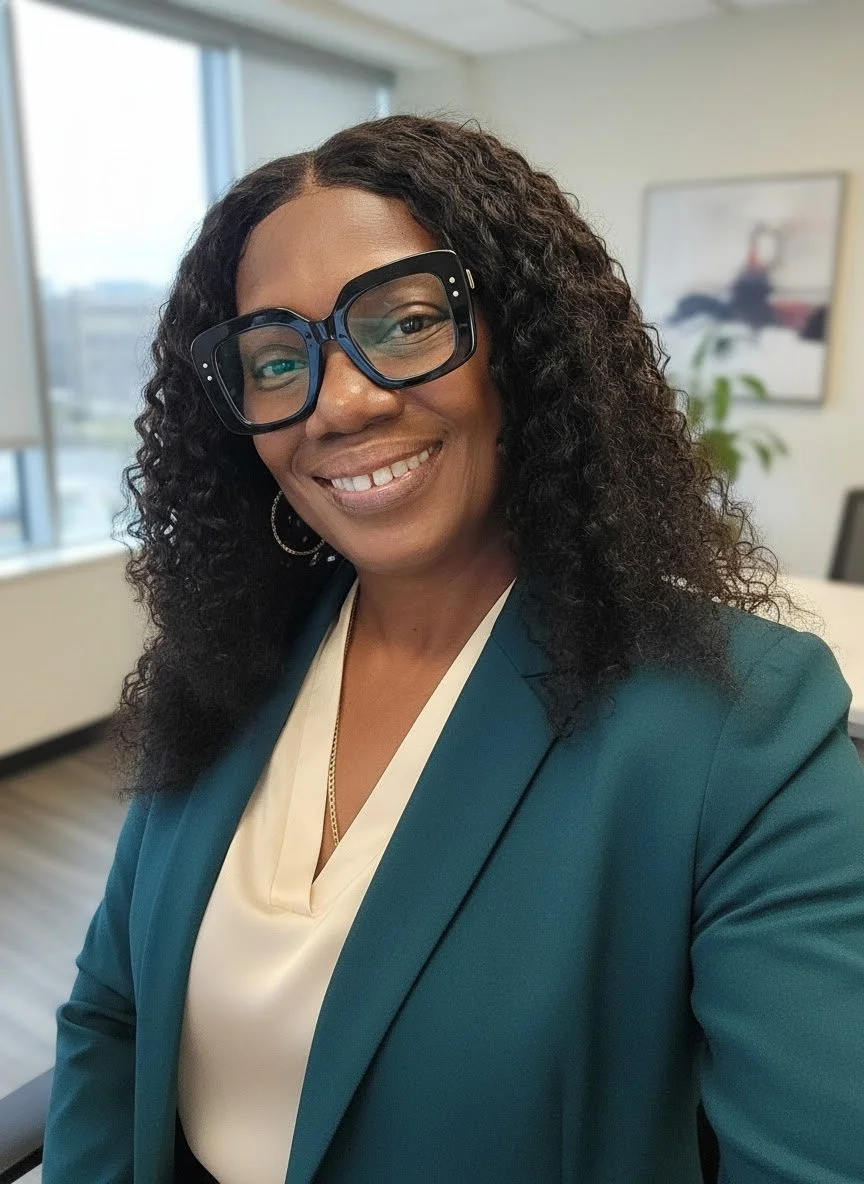 A woman with curly hair wearing large black glasses, a dark teal blazer, and a cream blouse, smiling in an office with windows and artwork in the background.