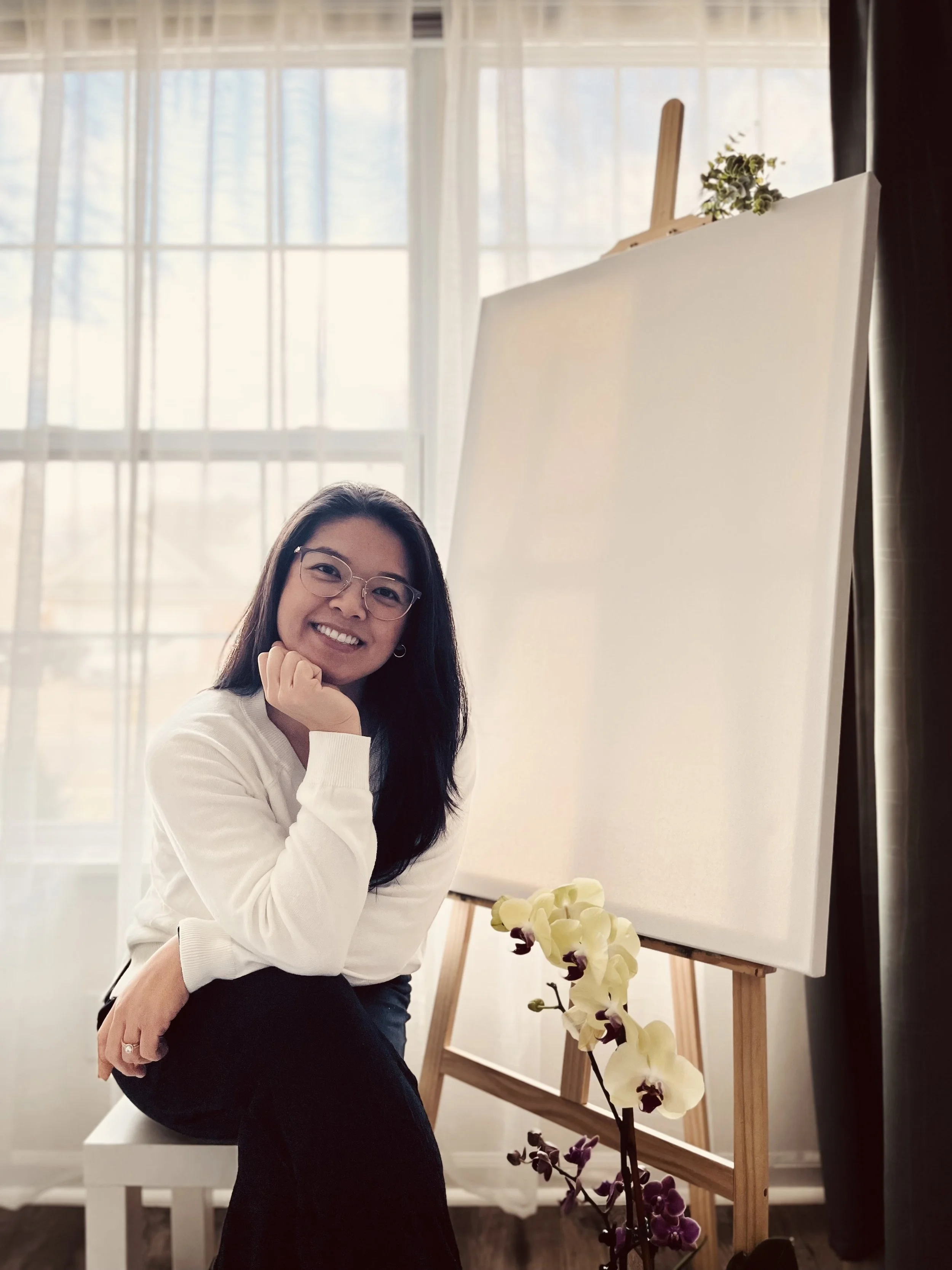 A woman with glasses, smiling and touching her chin, is seated near an empty easel with a plant in front of it, in a bright room with large windows and sheer curtains.