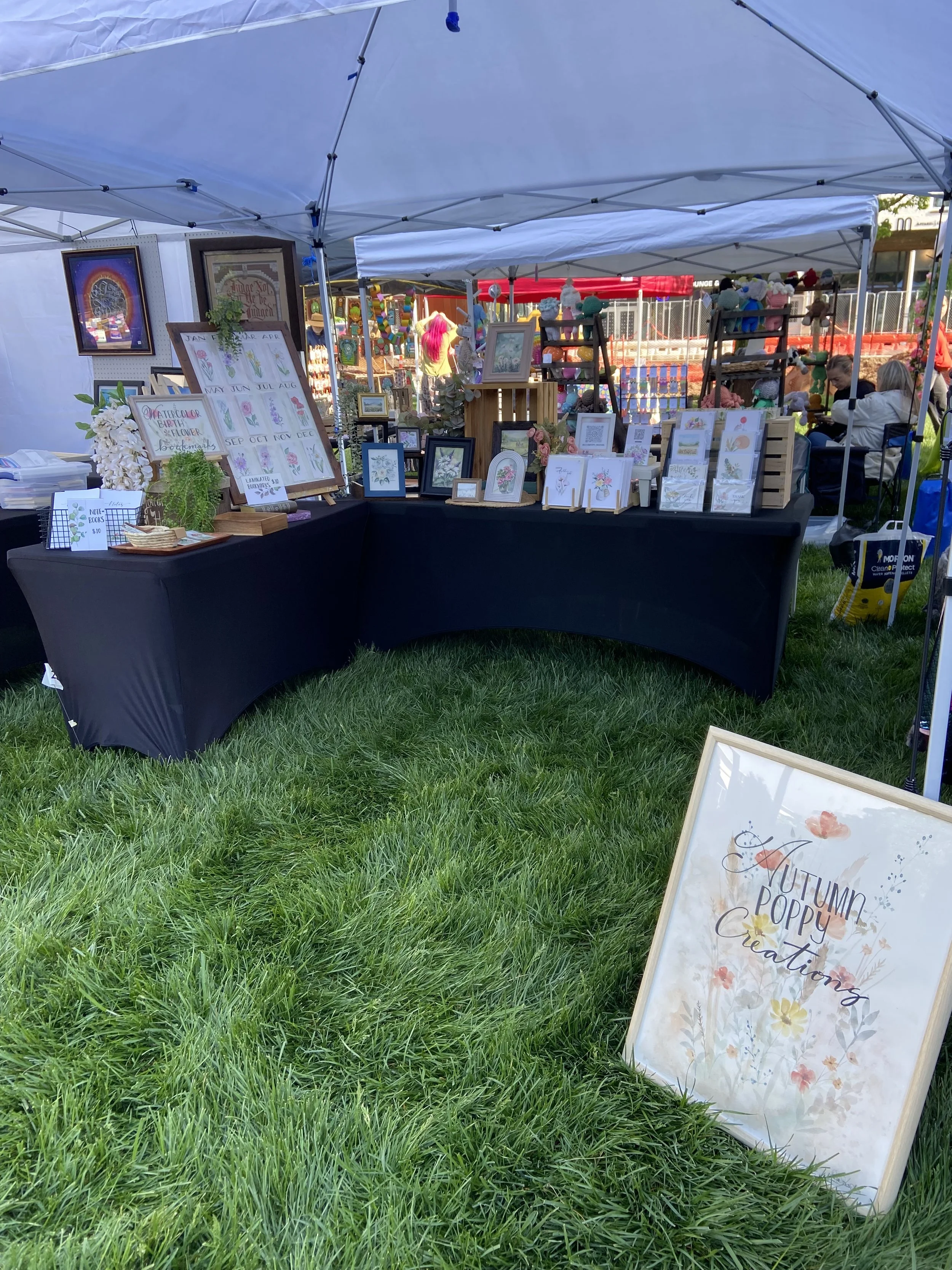 Outdoor art and craft booth at a fair or market, displaying framed artwork, greeting cards, and decorative items on tables and stands.