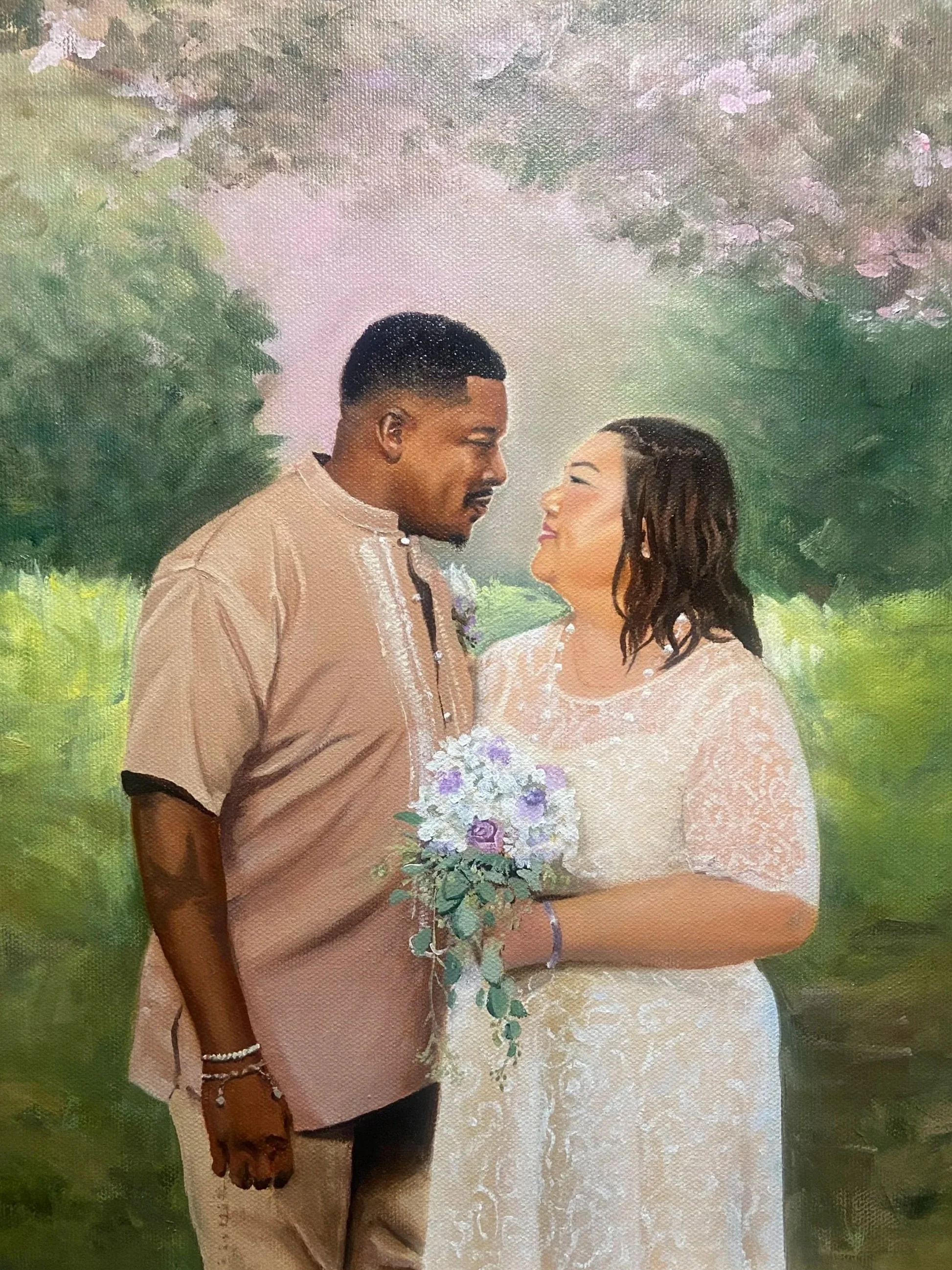 A couple standing outdoors, facing each other and smiling. The man is dressed in a beige shirt and black pants, and the woman is in a white lace dress. She holds a bouquet of white and purple flowers, and they are surrounded by green trees and foliag