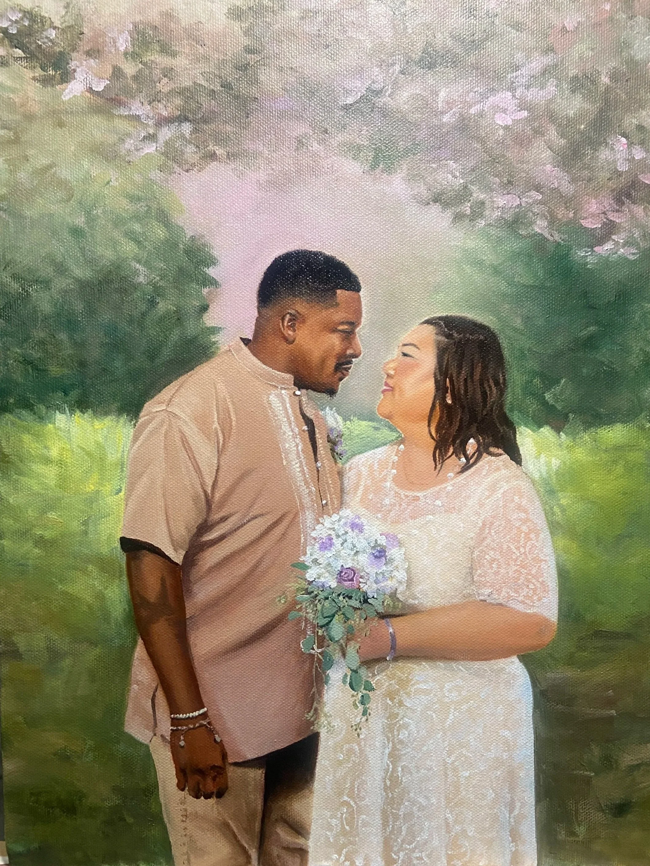 A couple stands closely together looking into each other's eyes outdoors, surrounded by lush greenery and pink flowering trees. The woman is wearing a white lace dress and holding a bouquet of white and purple flowers. The man is dressed in beige tra