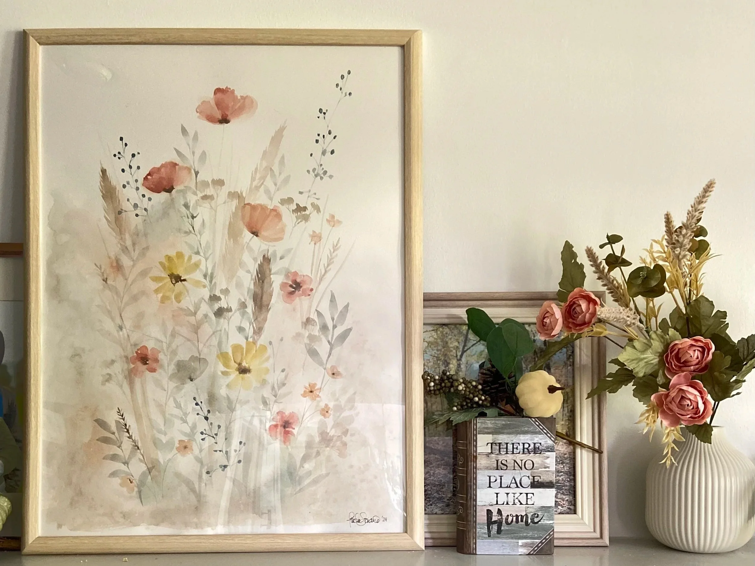 A framed watercolor floral painting on a wall next to a floral arrangement and a decorative box with the quote 'There is no place like home'