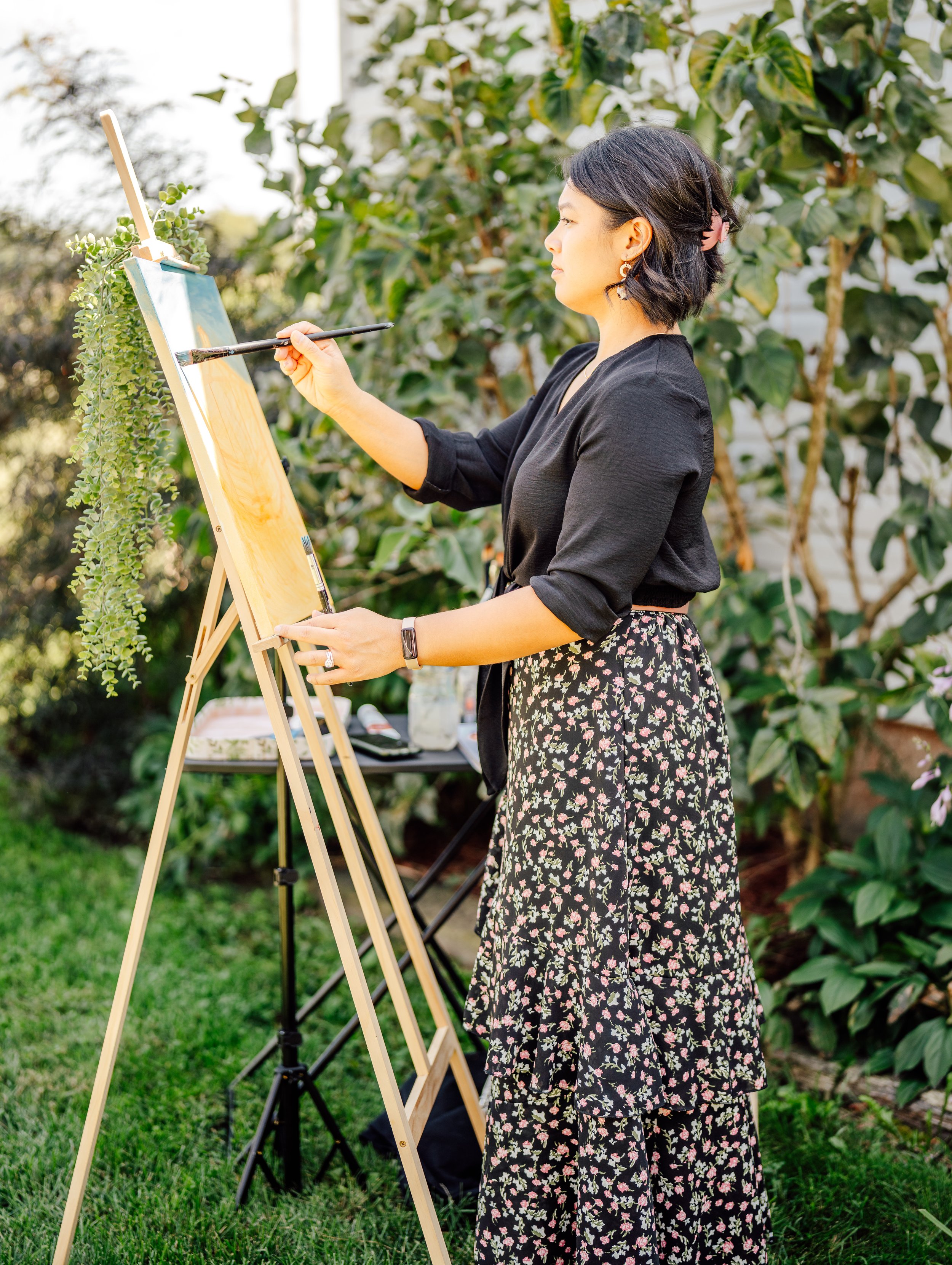 Woman wearing a black top and floral skirt painting on an easel outdoors with greenery in the background.