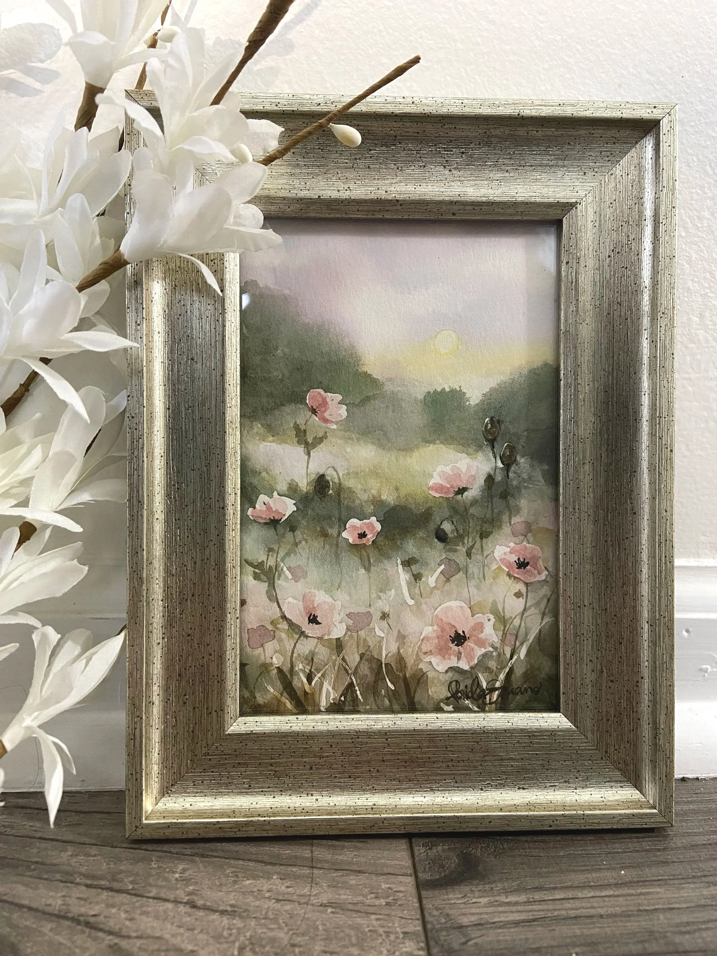A decorative framed watercolor painting of a field of pink poppies with a sunset or sunrise in the background, leaning against a wall with white lilies on the left side of the image.