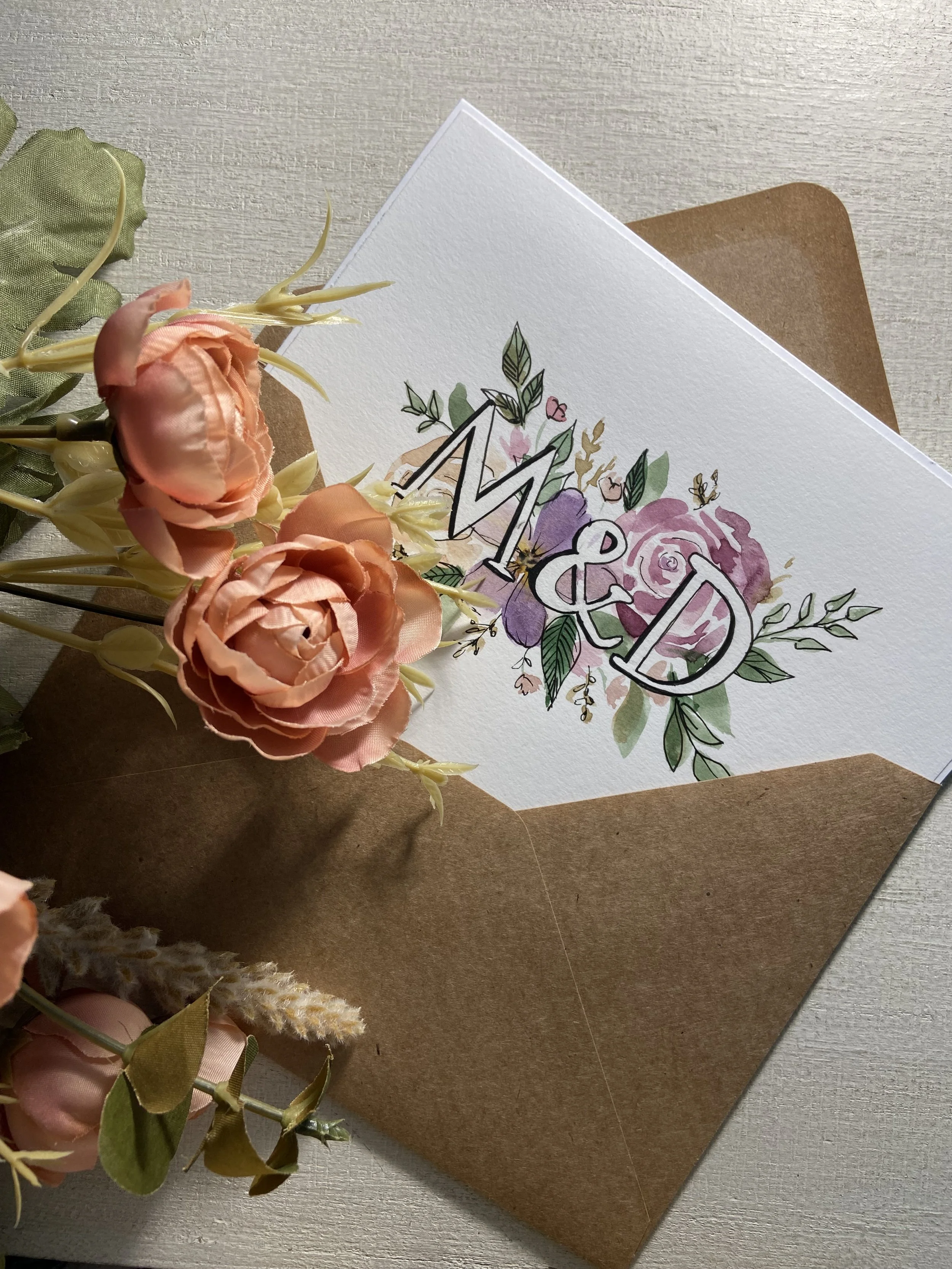 A floral greeting card with pink and purple flowers, green leaves, and the words 'Mr & Mrs' written on it, resting on a brown envelope and surrounded by peach-colored artificial roses and greenery.