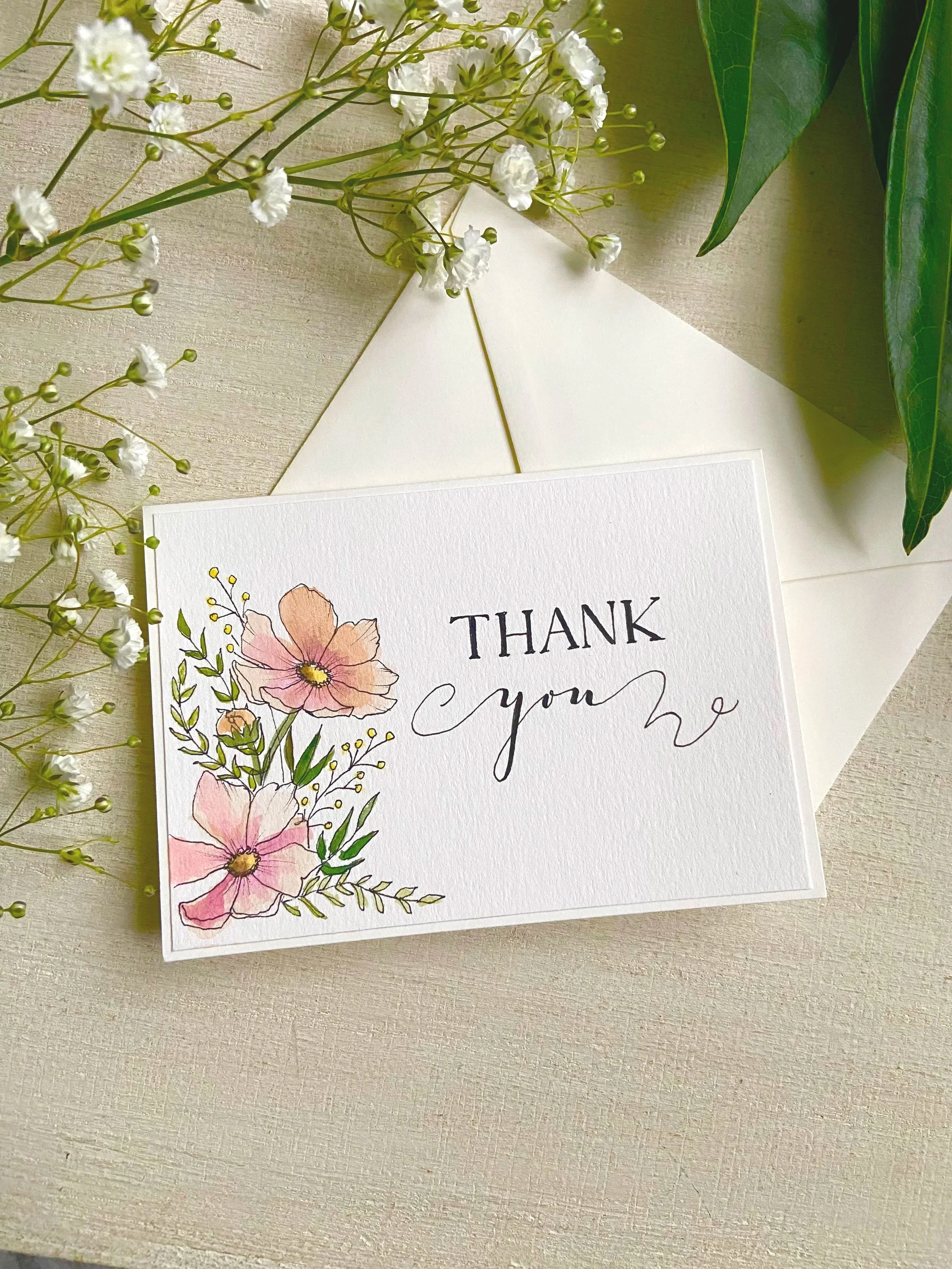 A thank you card with watercolor pink flowers and greenery, resting on a light-colored textured surface, surrounded by white baby's breath flowers and green leaves.
