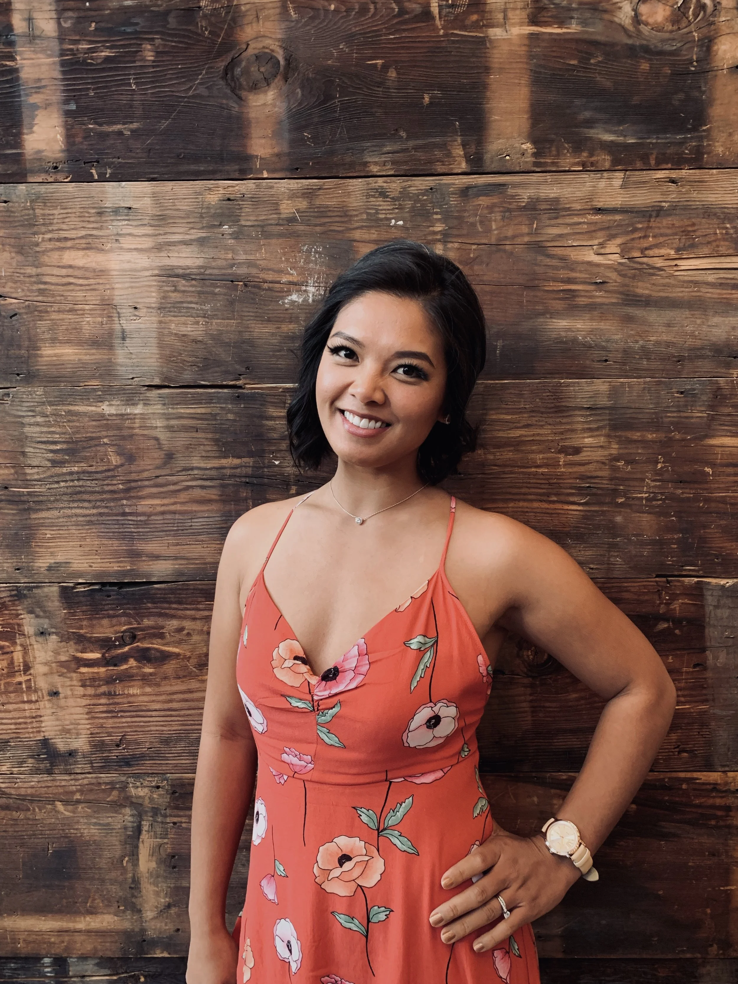 A woman with black, wavy, shoulder-length hair, wearing a coral-colored dress with floral patterns and thin straps, standing in front of a wooden wall with horizontal planks. She is smiling, wearing a necklace, a watch, and a ring, with her left hand on her hip.
