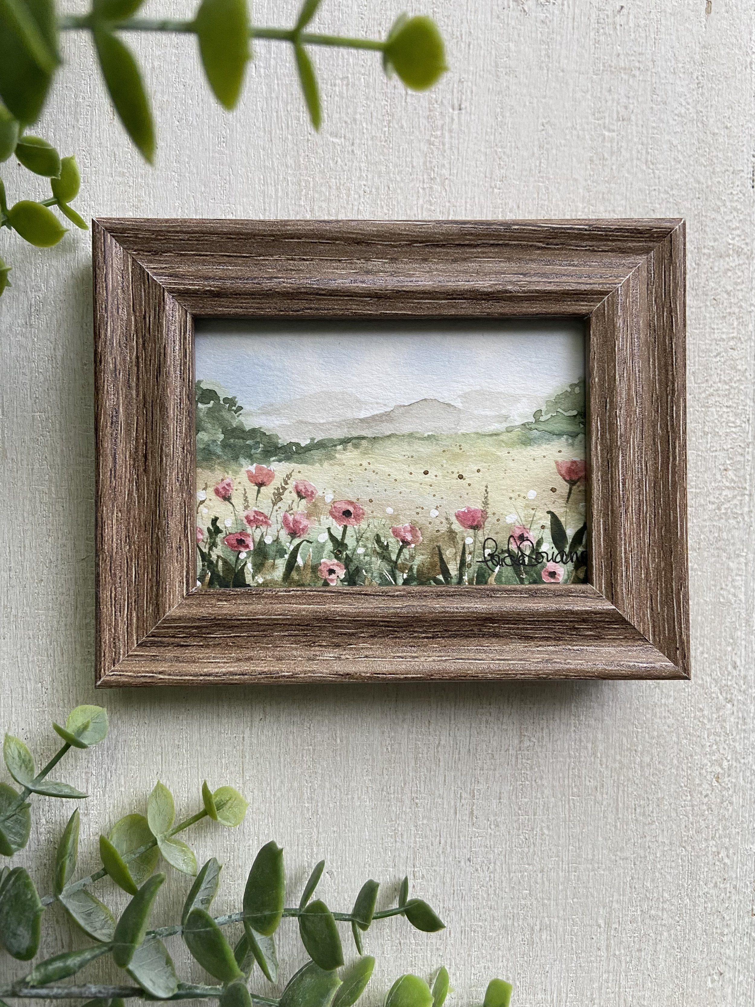 Close-up view of a framed watercolor painting of a landscape with pink flowers in the foreground, green trees, and distant mountains. The frame is wooden with a textured finish. Green leaves are partially visible on the left side of the image, and the background surface is light-colored textured wall.