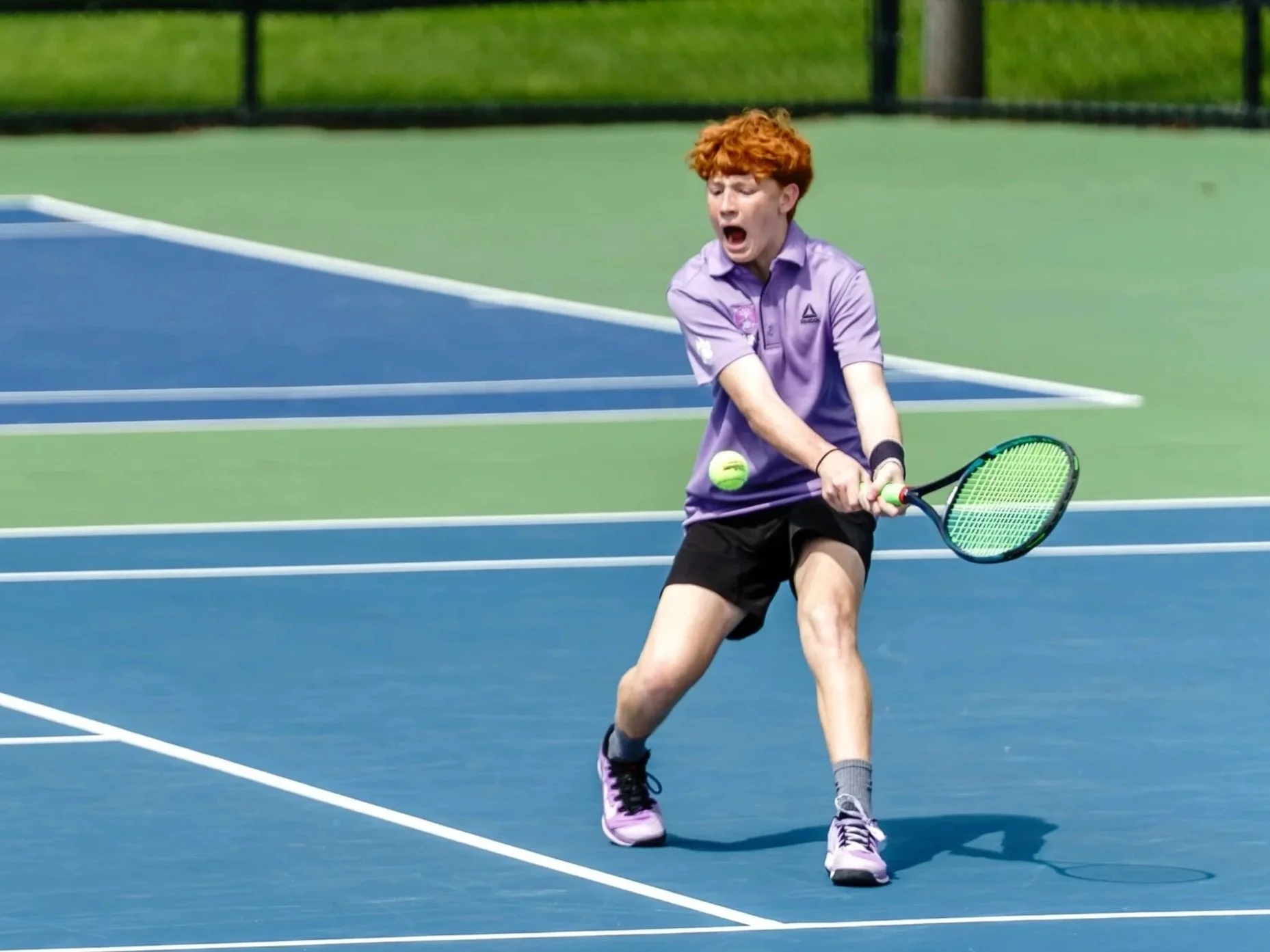 A young person with curly red hair playing tennis on a blue court, wearing a purple shirt, black shorts, and purple tennis shoes, with a tennis ball about to be hit with a racket.