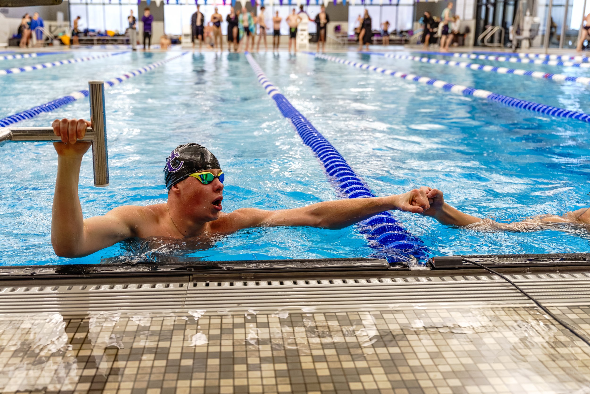 Swimmer holding onto the edge of an indoor swimming pool, wearing a black swim cap and goggles, with swim lane ropes and people in swimsuits in the background.