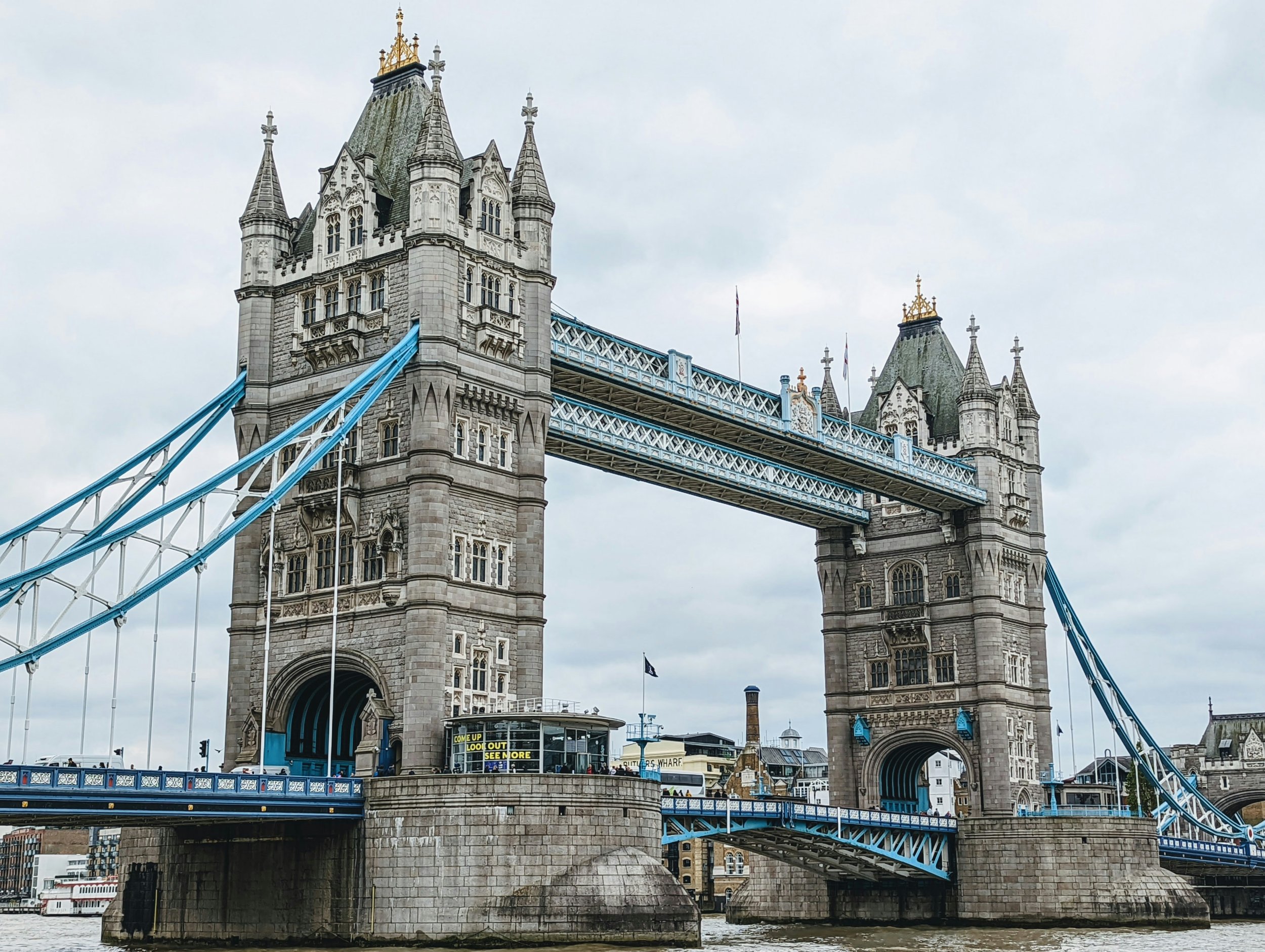 London Tower Bridge with its towers, suspension cables, and bascule sections over the River Thames, with cloudy sky in the background.