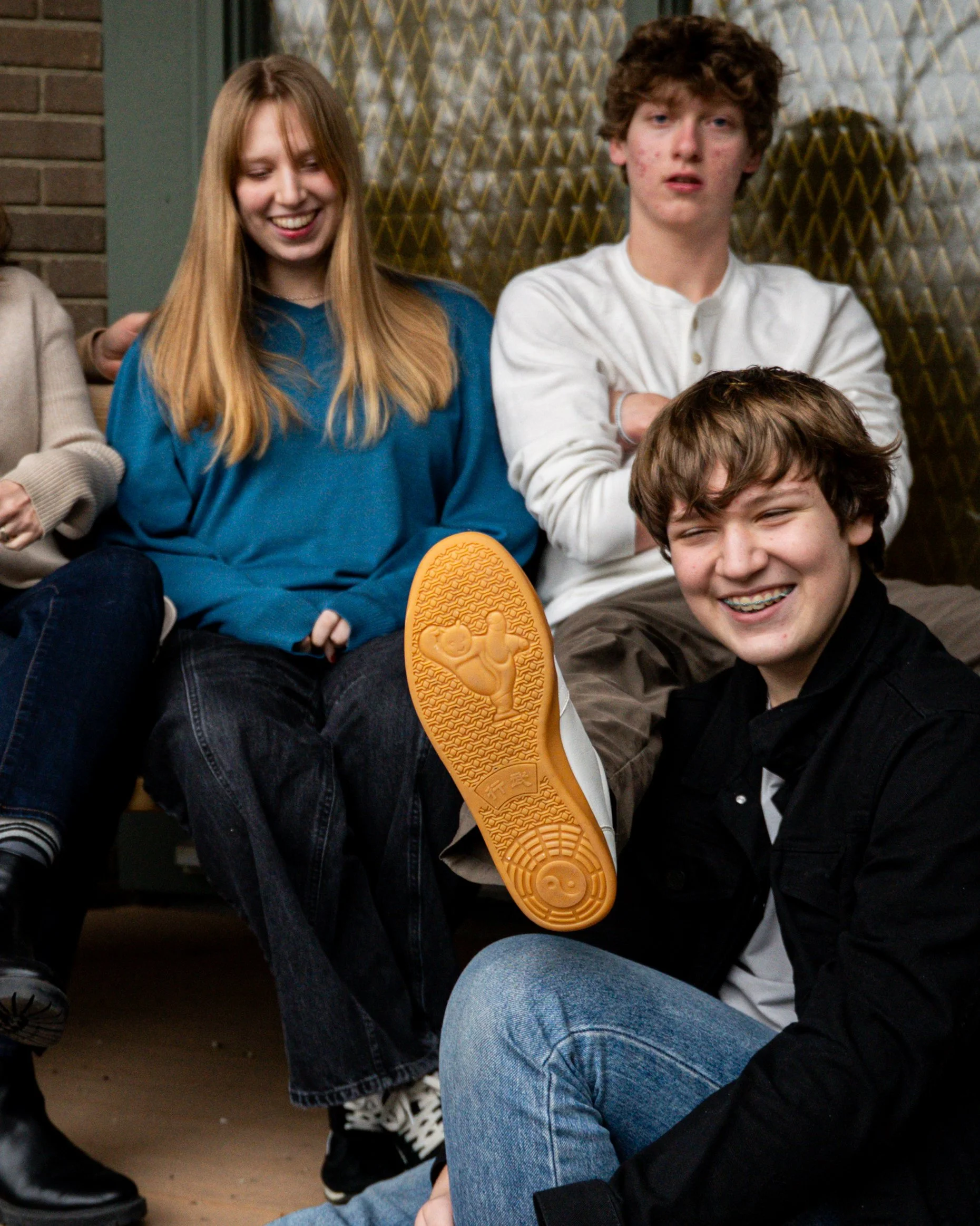 Three teenagers sitting on the floor, smiling. One girl in a blue shirt is holding a yellow shoe with a cartoon figure on the sole. Two boys are nearby, one with braces and one with curly hair.