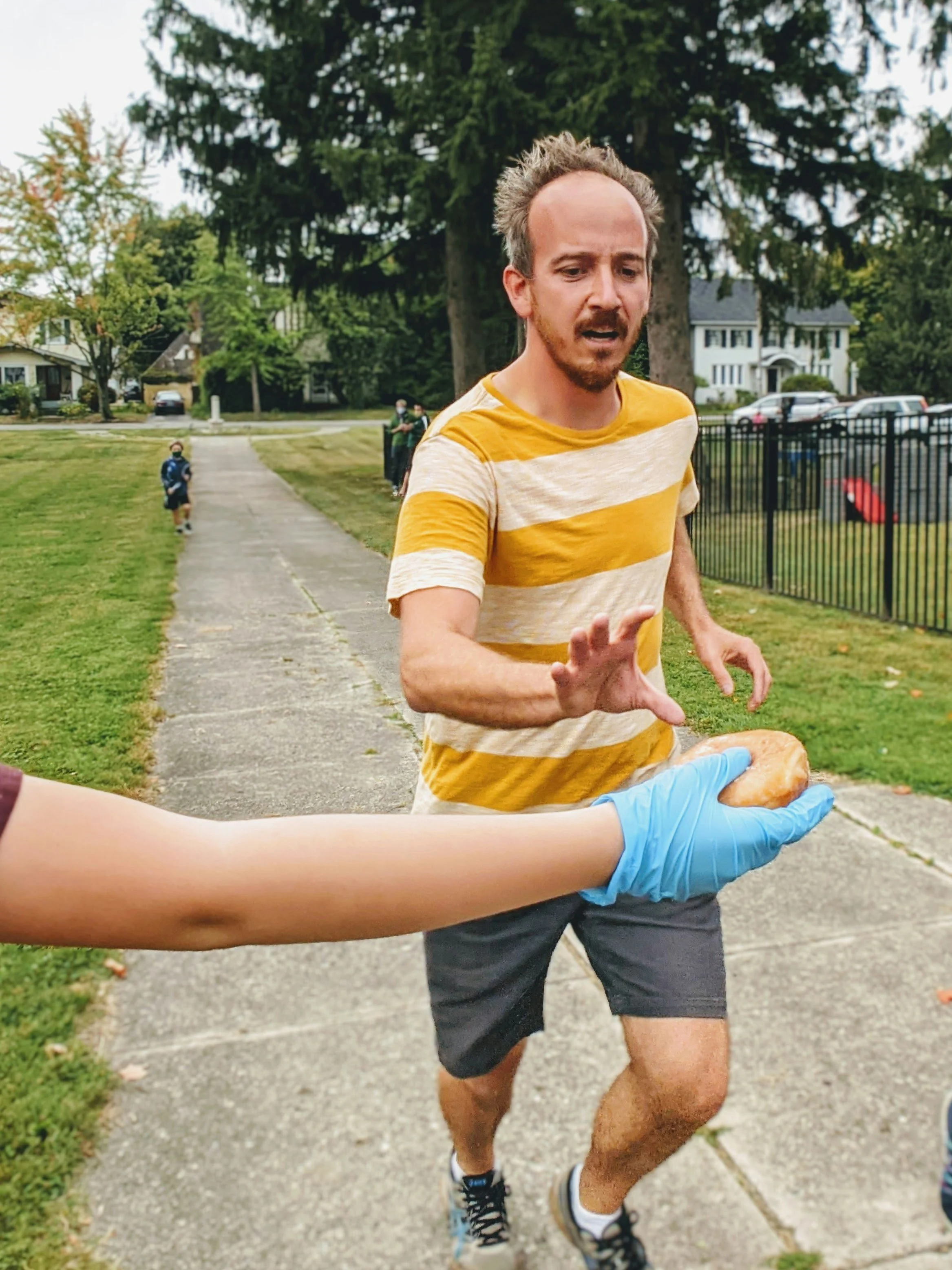 A man running outdoors on a sidewalk, wearing a yellow and white striped T-shirt, black shorts, and black running shoes with white socks, while a person in a blue glove hands him a donut.