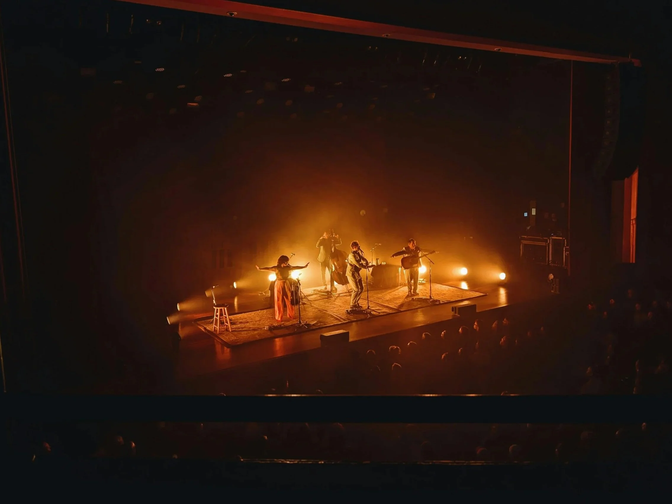 A stage performance with five musicians in a dark theater, illuminated by warm stage lights, featuring two guitar players, a drummer, and two singers, all in casual attire.