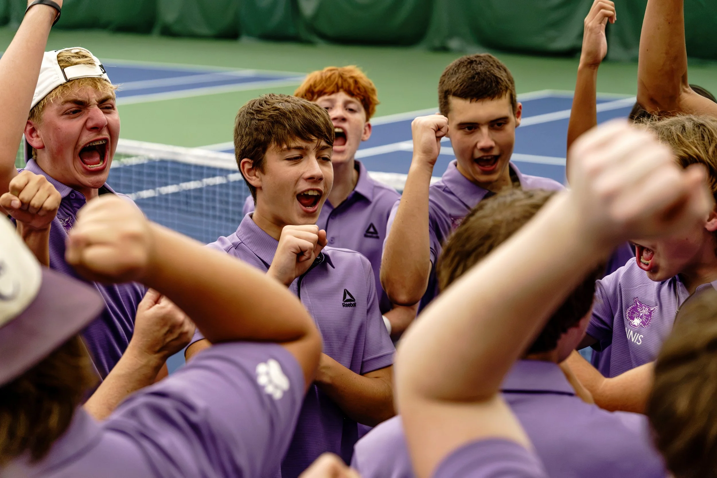 Group of young athletes in purple uniforms celebrating with raised fists on a tennis court.