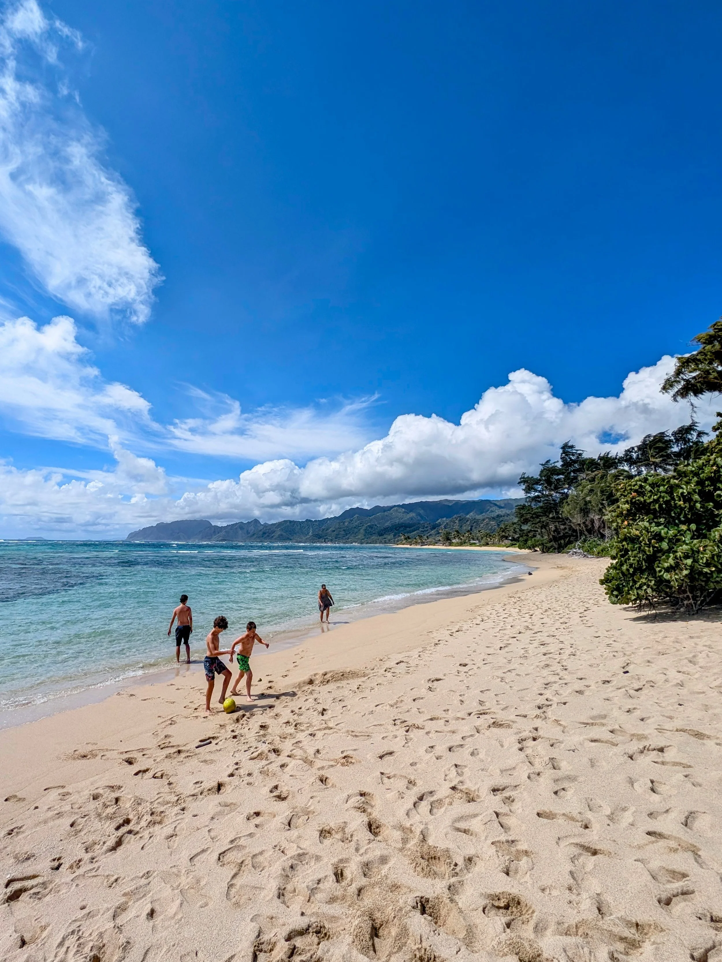 People playing soccer on a sandy beach near the ocean, with a backdrop of mountains, trees, and a partly cloudy sky.