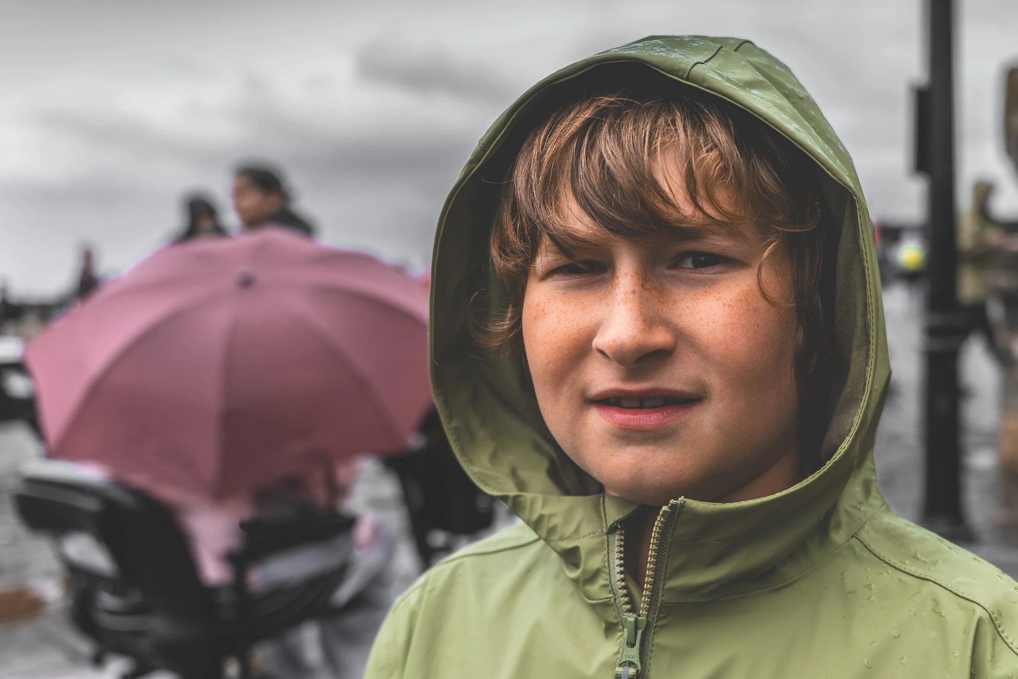 A young boy with brown hair, freckles, and blue eyes, wearing a green rain jacket with the hood up, standing in the rain. In the background, there are blurred figures and an umbrella on a cloudy day.