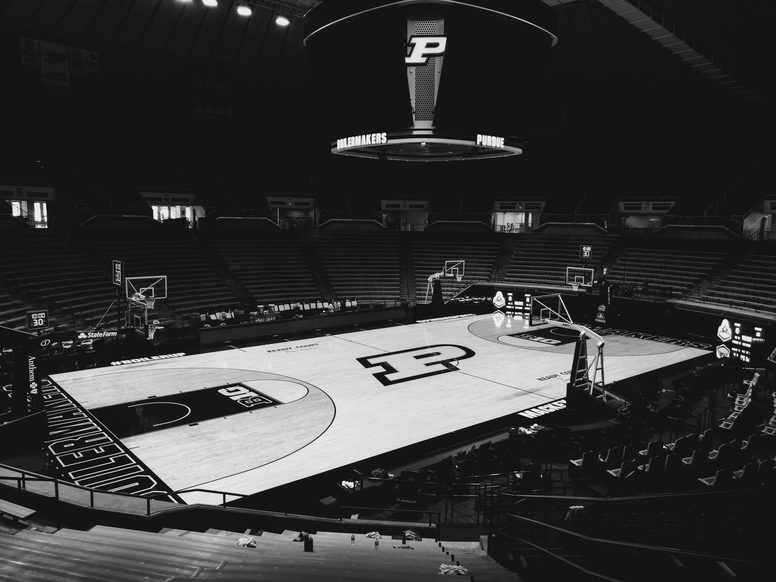 An empty basketball arena with a hardwood court in the center, featuring the Purdue Boilermakers logo and branding, basket hoops, digital scoreboards, and rows of seating surrounding the court.