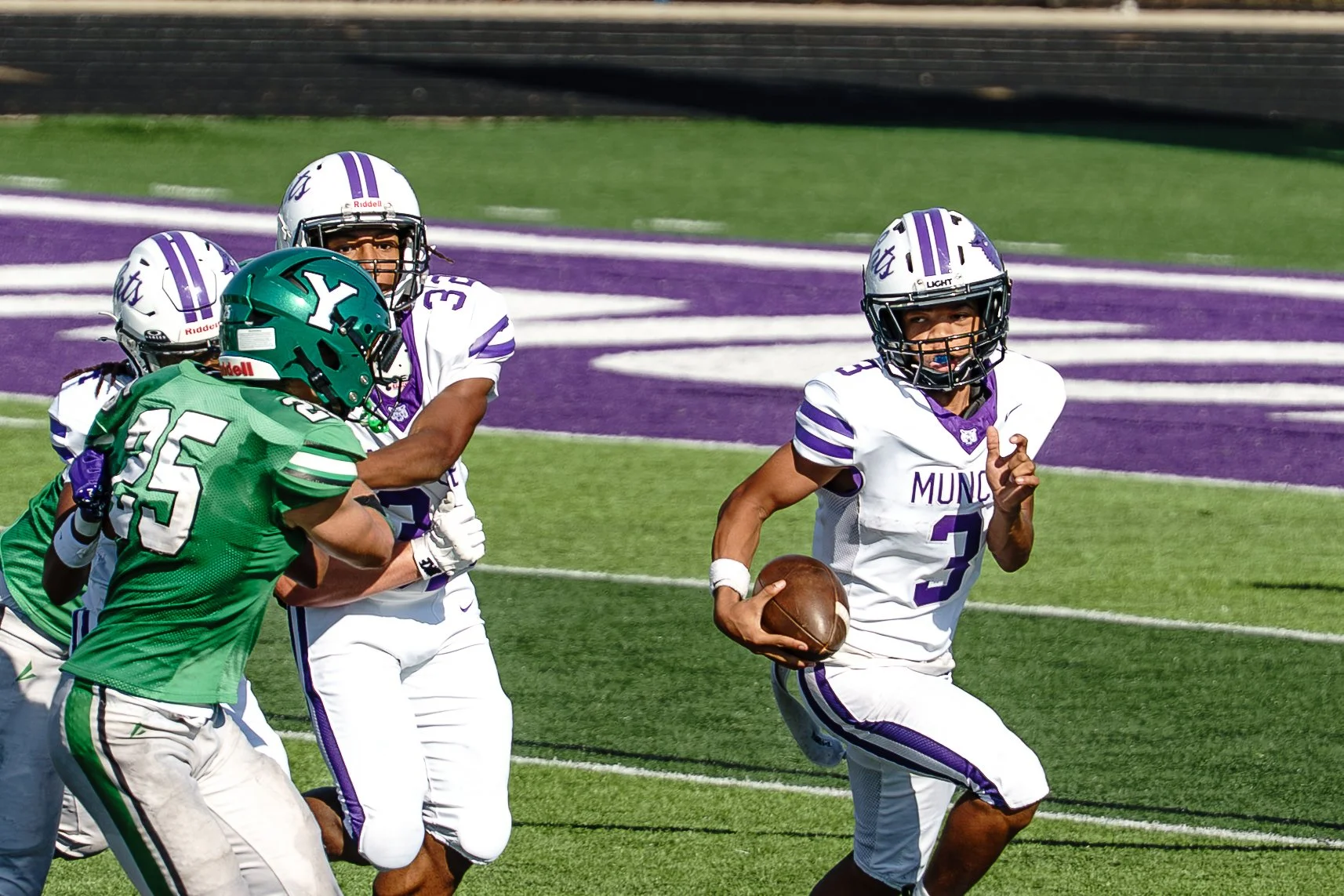 A football game with players from two teams: one team in white jerseys with purple accents, and the other in green jerseys. The player in white, wearing number 3, is running with the football, while players from the opposing team try to tackle him, on a field with purple and green markings.