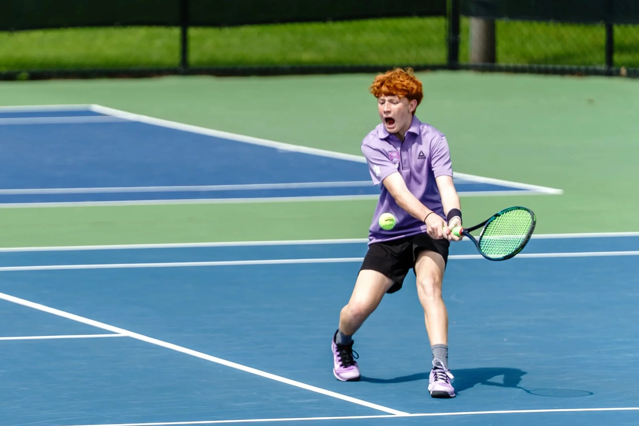 A young person with red curly hair playing tennis on an outdoor court, preparing to hit a tennis ball with a racquet.