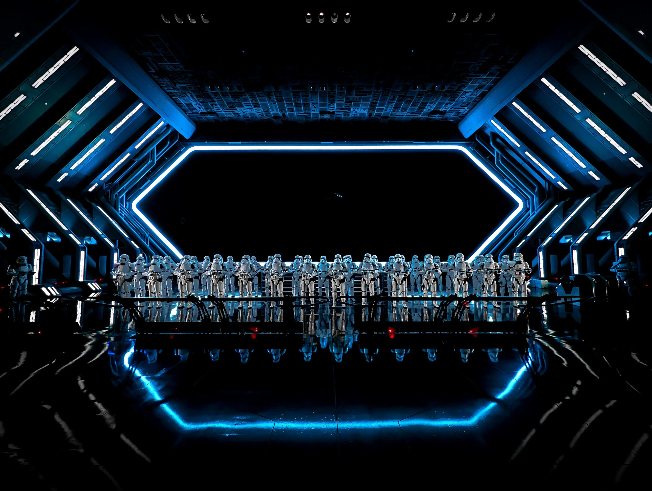A futuristic sci-fi scene with a row of white stormtroopers from Star Wars standing inside a sleek, illuminated spaceship or space station. The space features a high-tech, blue neon-lit geometric design with reflective surfaces.