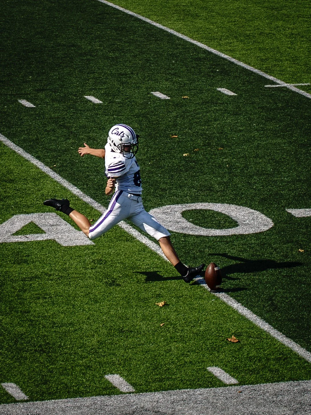 A young football player in a white and purple uniform runs on a green football field, just as they kick the football from the ground.