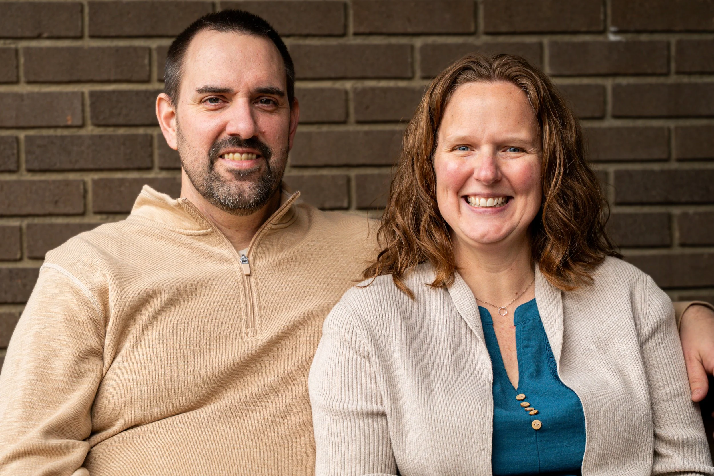 A man and a woman standing close together against a brick wall, smiling at the camera.