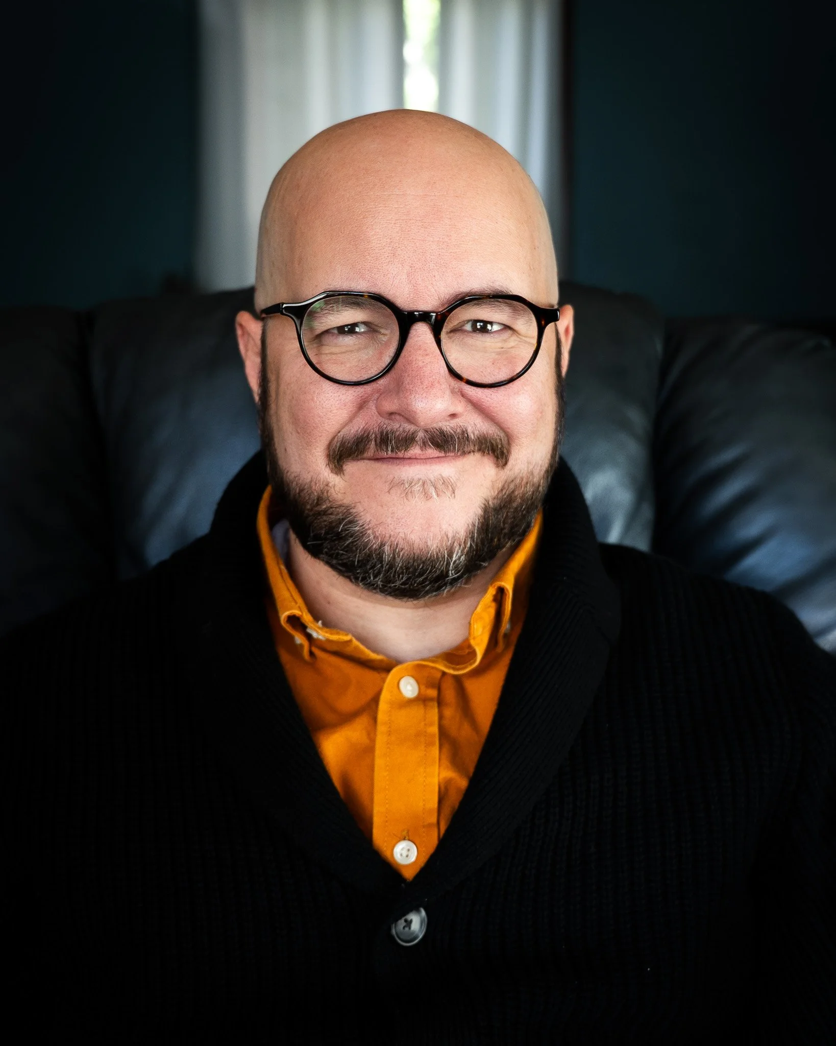 A man with a bald head, glasses, and a beard smiles while sitting on a black armchair. He is wearing a black sweater over an orange collared shirt with white buttons.