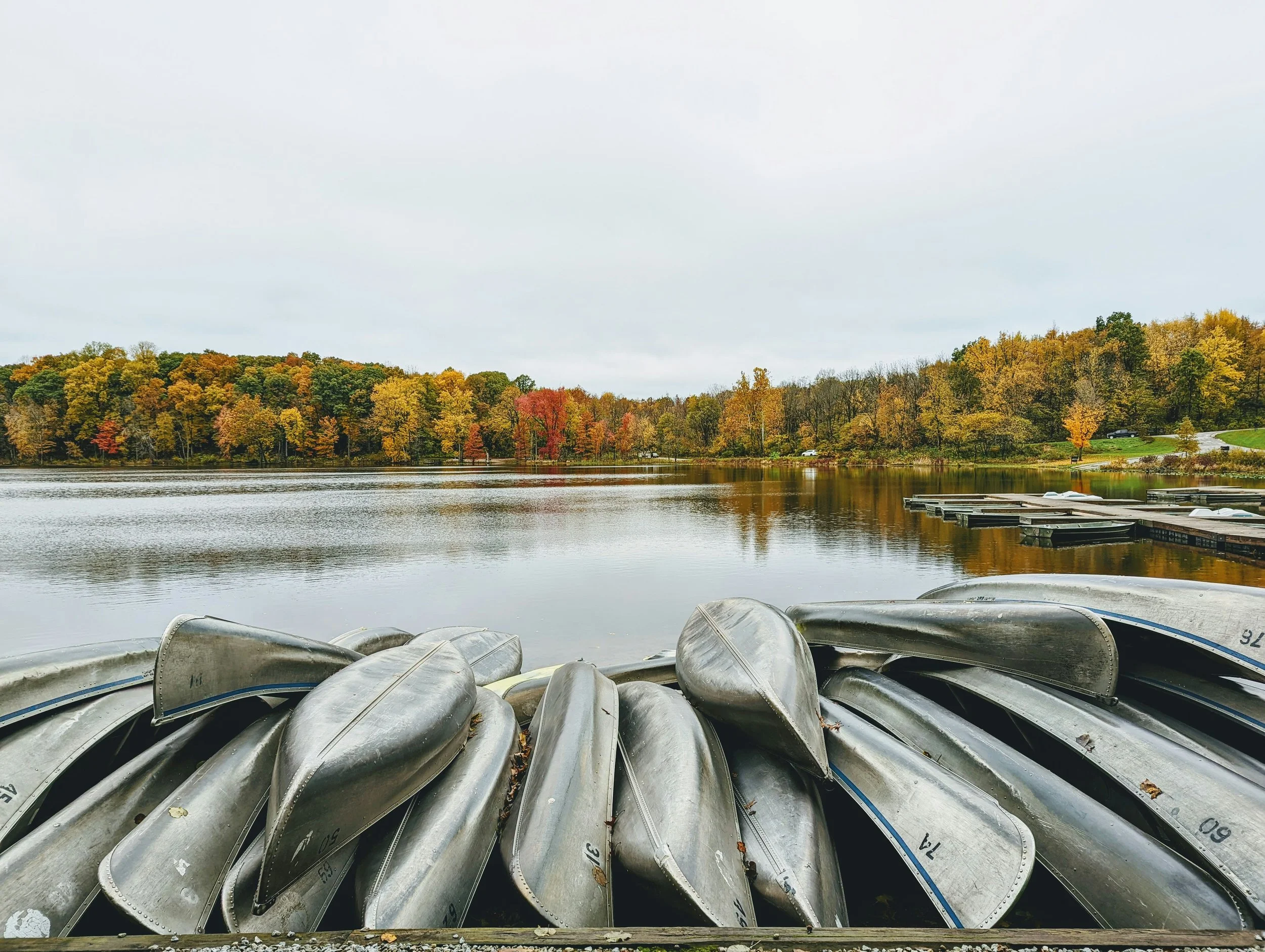 A row of aluminum canoes stacked upside down on a dock by a river, with trees displaying fall foliage in the background and a cloudy sky overhead.