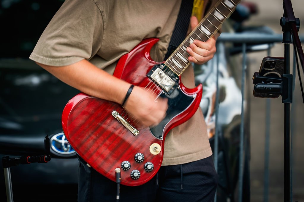 Person playing a red electric guitar during an outdoor performance with a microphone stand nearby.