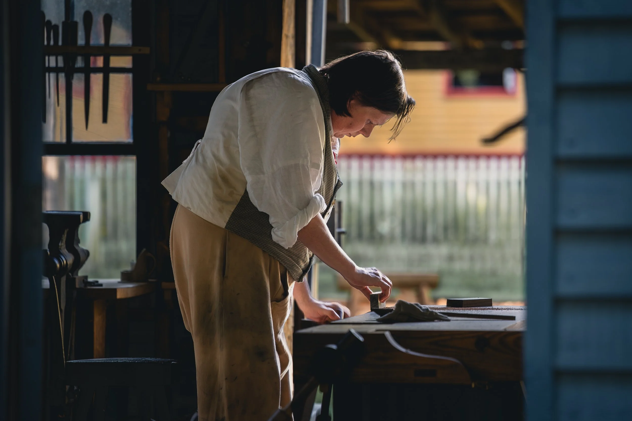 A woman with dark hair, wearing a beige apron and a white shirt, is leaning over a workbench in a wooden workshop, engaged in a woodworking project.
