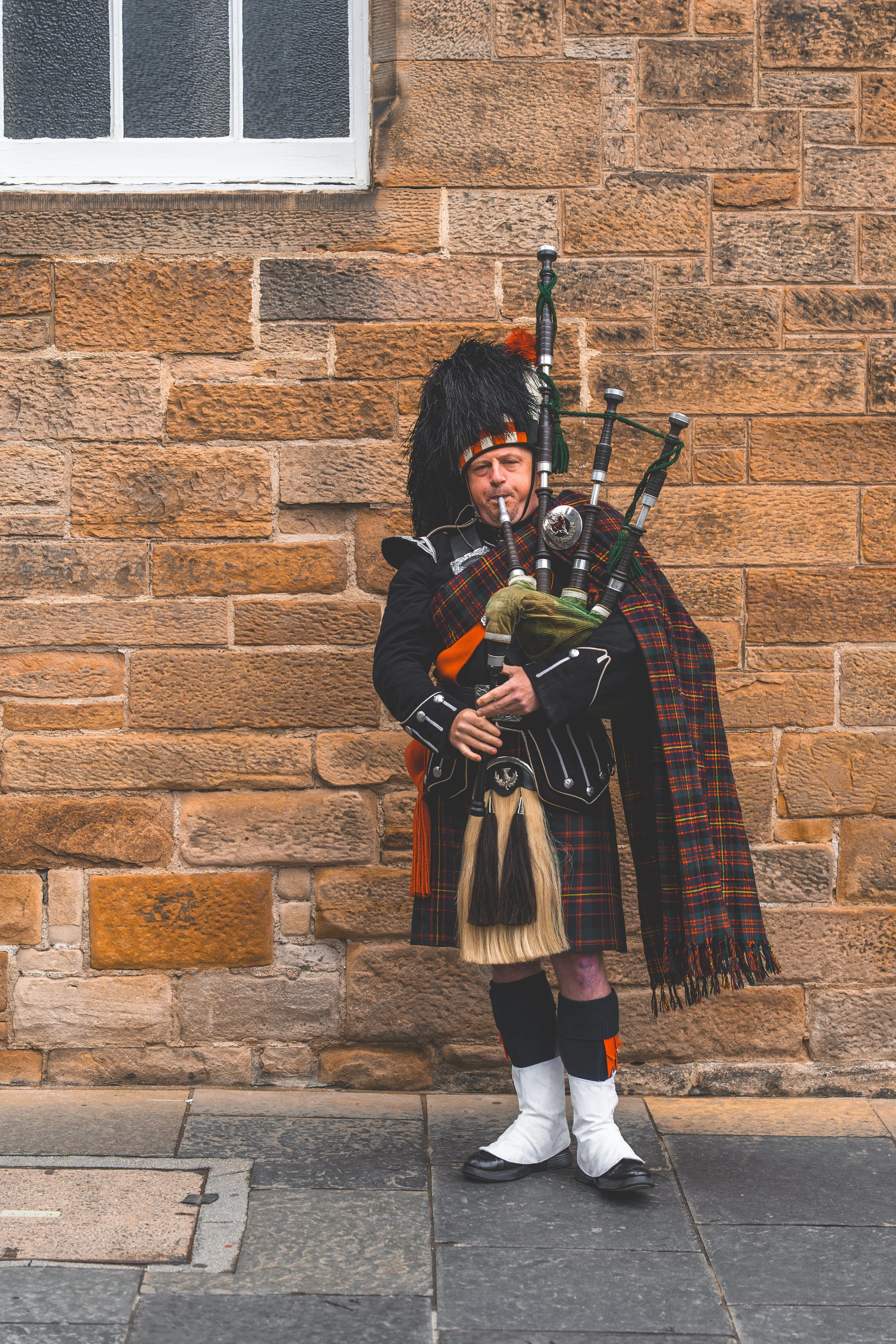 A man dressed in traditional Scottish Highland attire playing bagpipes against a brick wall background.