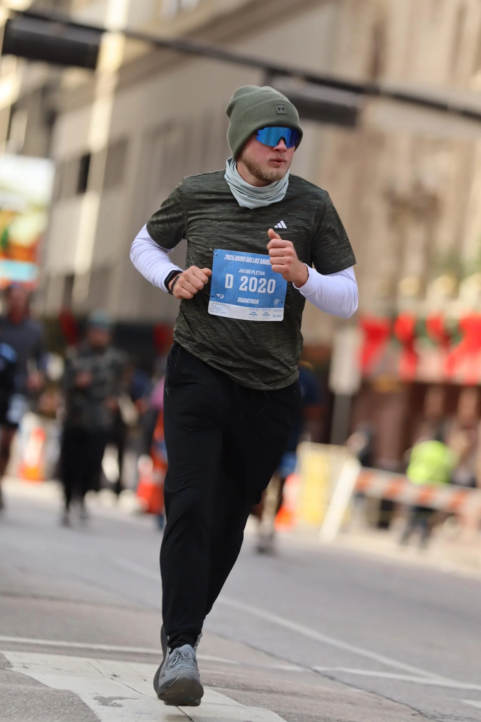 A man running in a marathon wearing a gray beanie, blue glasses, a gray Adidas shirt, black pants, and white sneakers, with a race bib numbered D 2020.