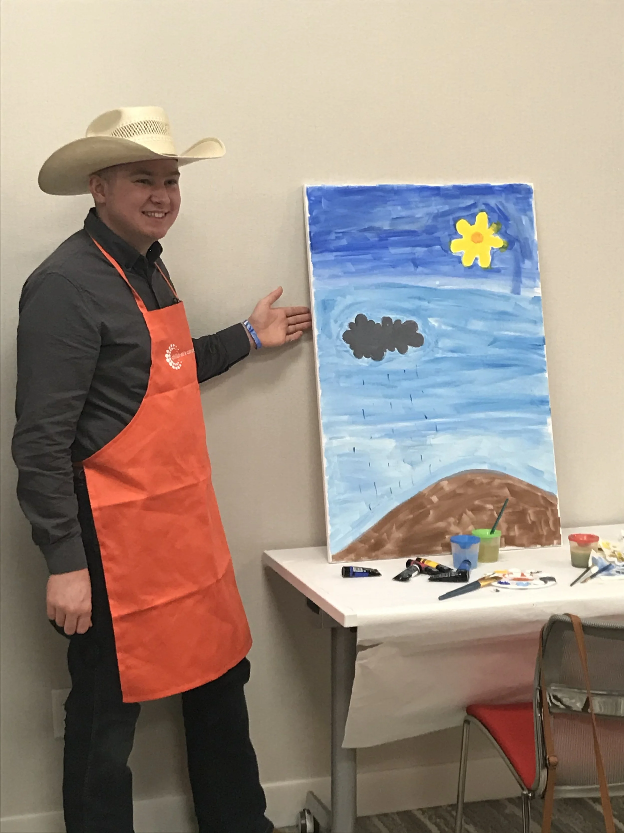 A young man wearing a cowboy hat and an orange apron, standing next to a painting on an easel depicting a blue sky with a yellow flower, a gray cloud, and brown hills. There are art supplies on the table nearby.