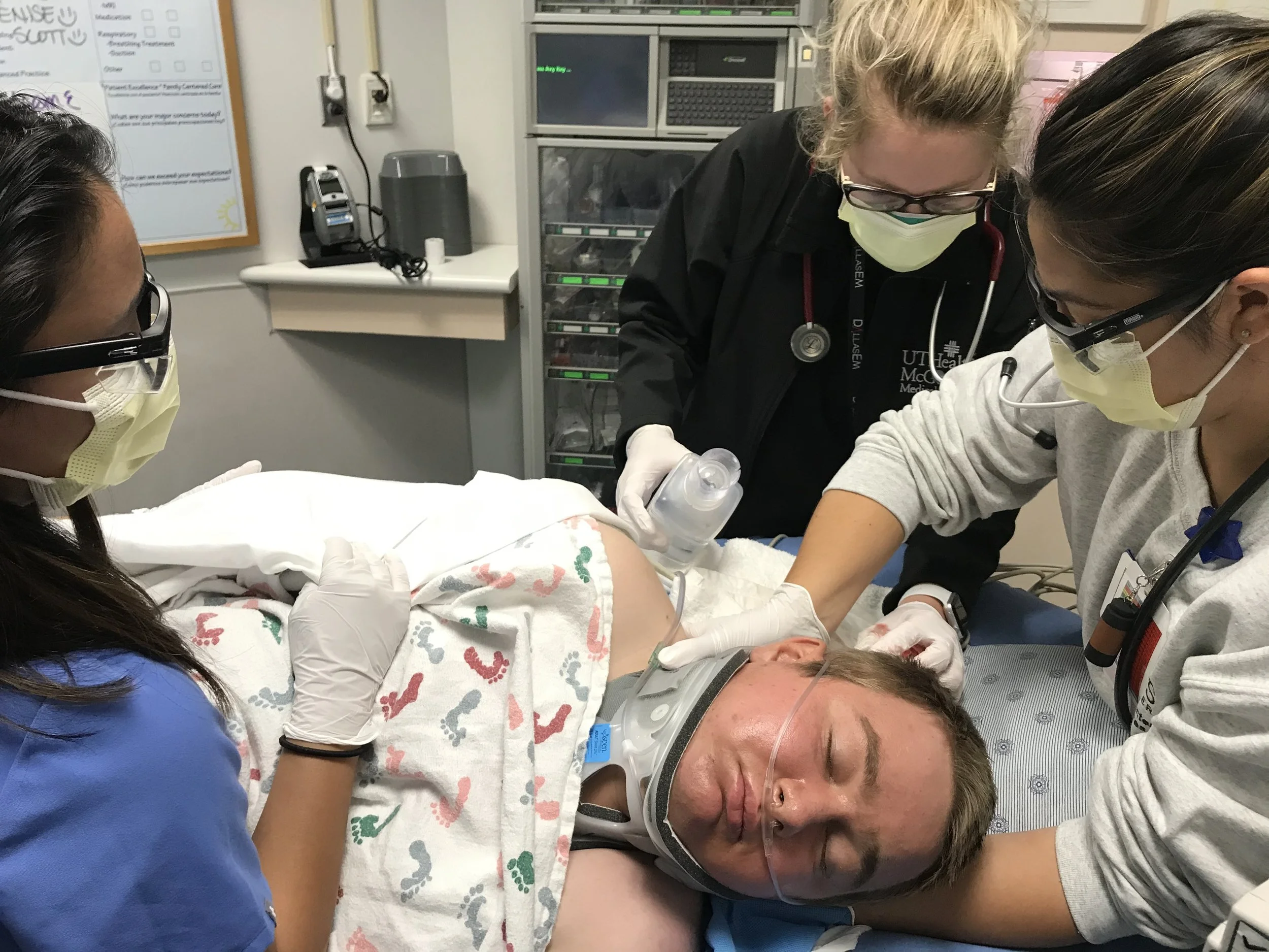Medical team attending to a patient in a hospital room, with healthcare professionals wearing masks and gloves, and various medical equipment visible in the background.