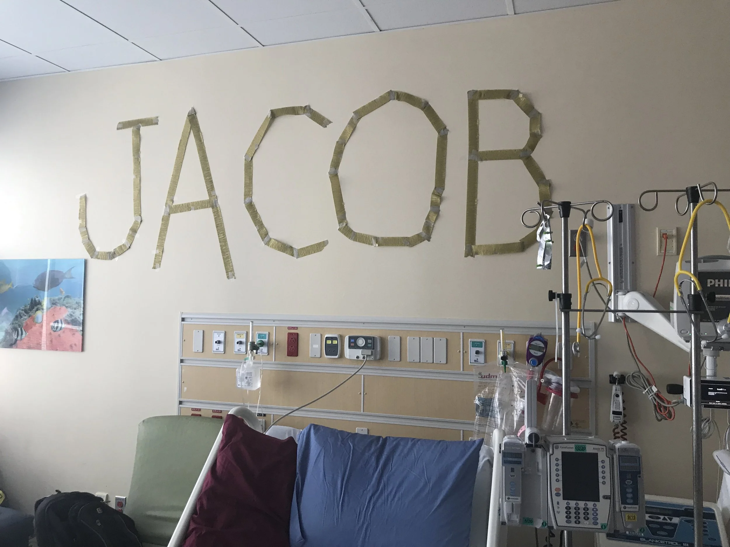 Hospital room with medical equipment and a hospital bed, wall with medical outlets, and the word 'TACO' spelled out on the wall with tape.