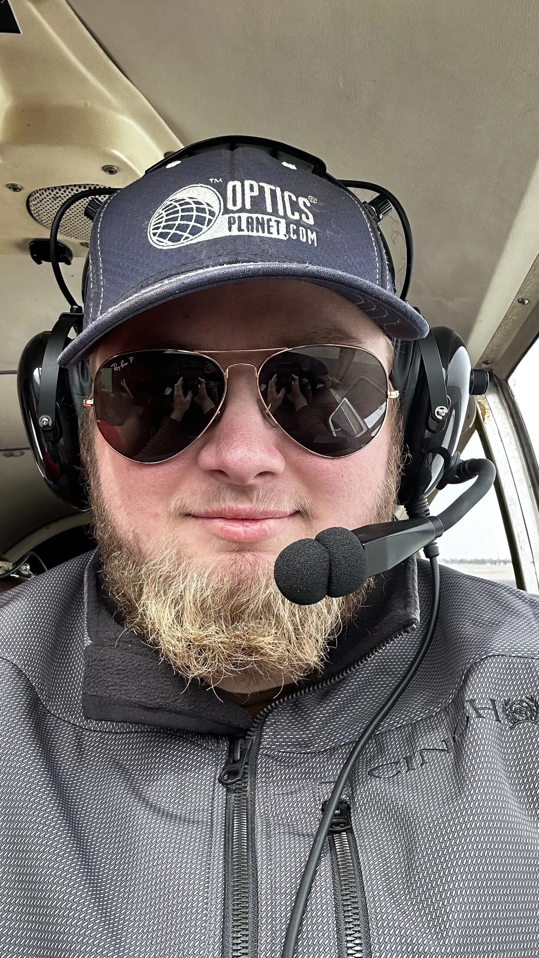 A man with a beard wearing sunglasses, a headset, a cap with 'OpticsPlanet.com' logo, and a gray jacket, smiling inside an aircraft.