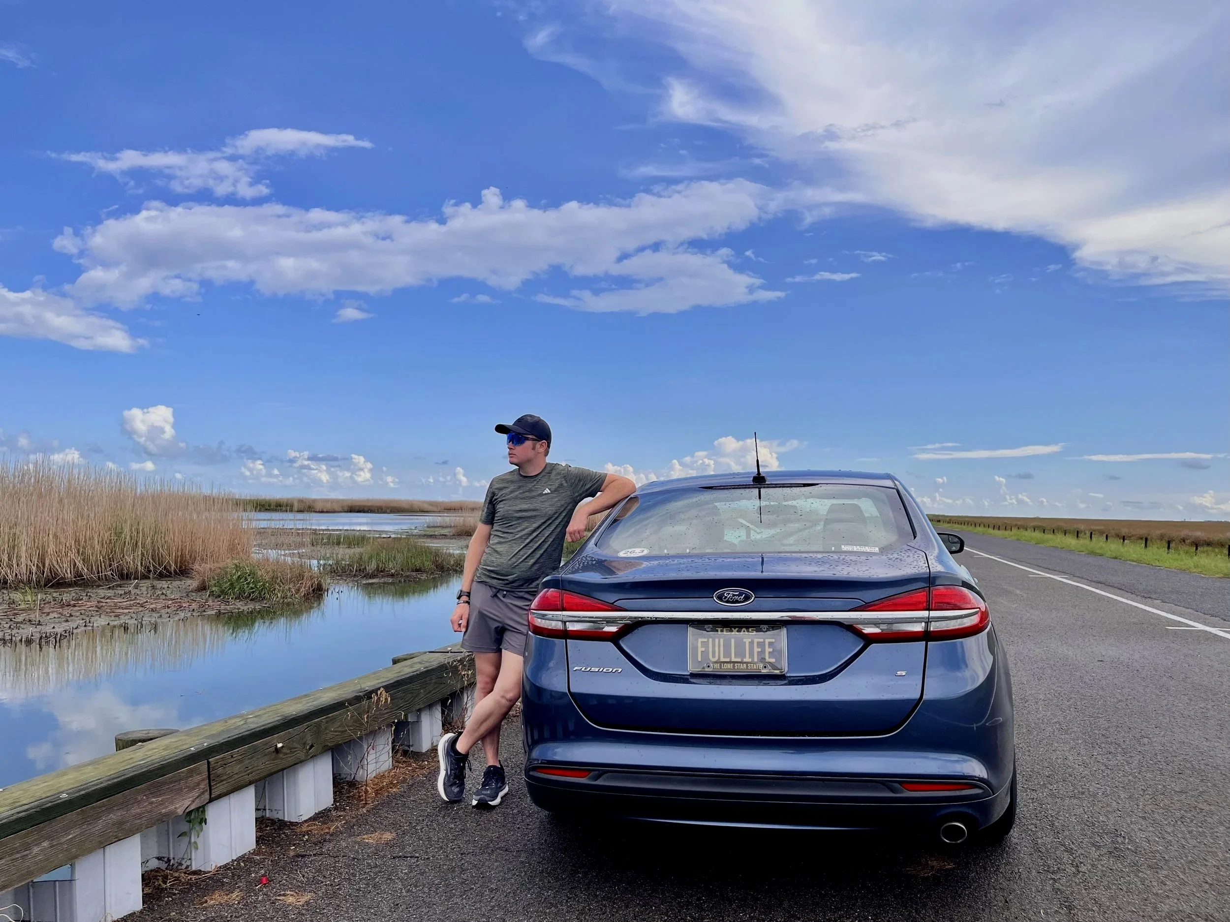 A man in athletic clothing and sunglasses leaning against a black Ford Fusion car with Texas license plate reading "FULL LIFE" parked beside a wooden guardrail near a marshland with tall grass and water. The sky is blue with scattered clouds.