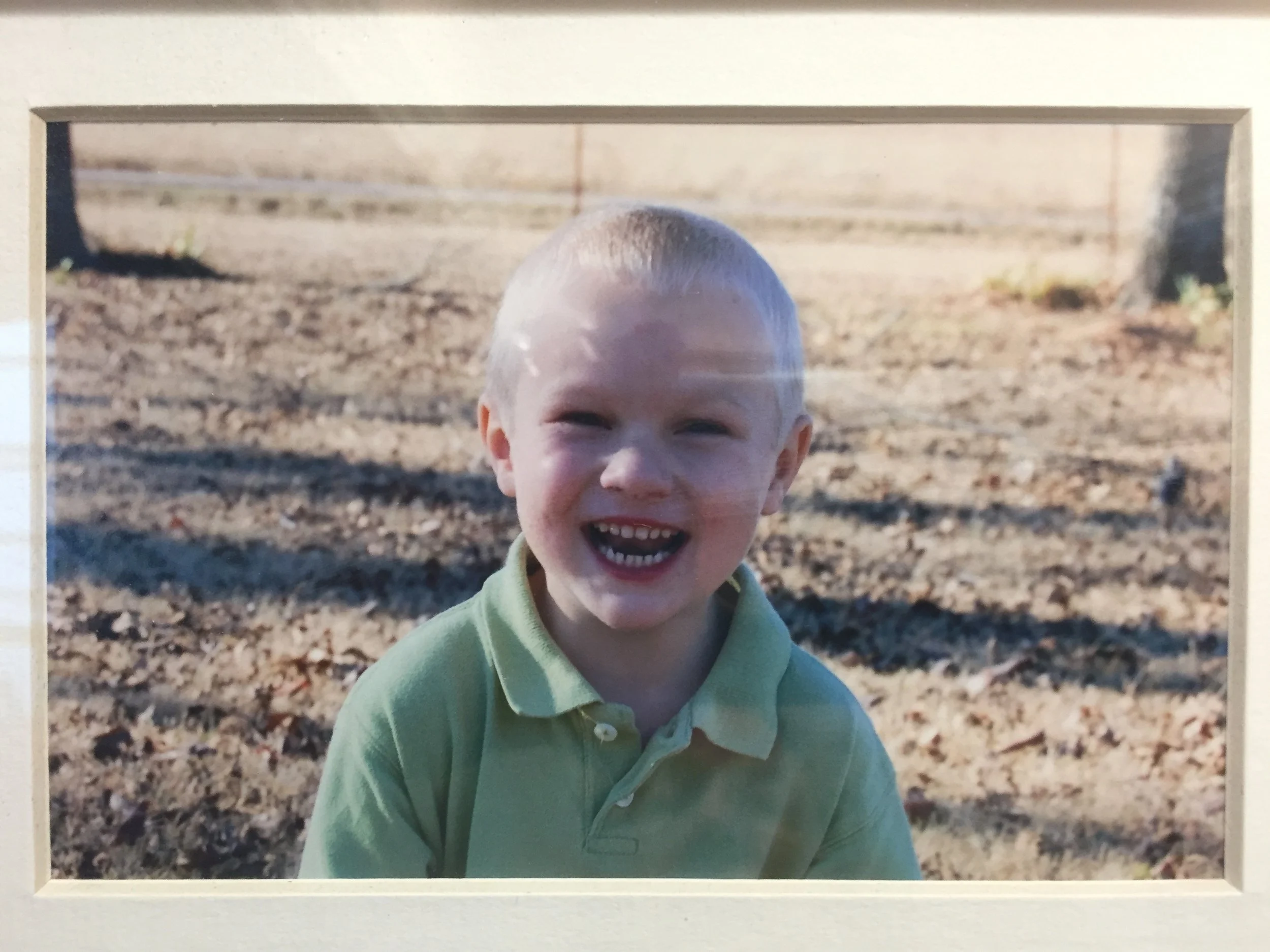 A smiling young boy with light hair and a green collared shirt standing outdoors on a sunny day, with a blurred background of bare ground and some sparse vegetation.