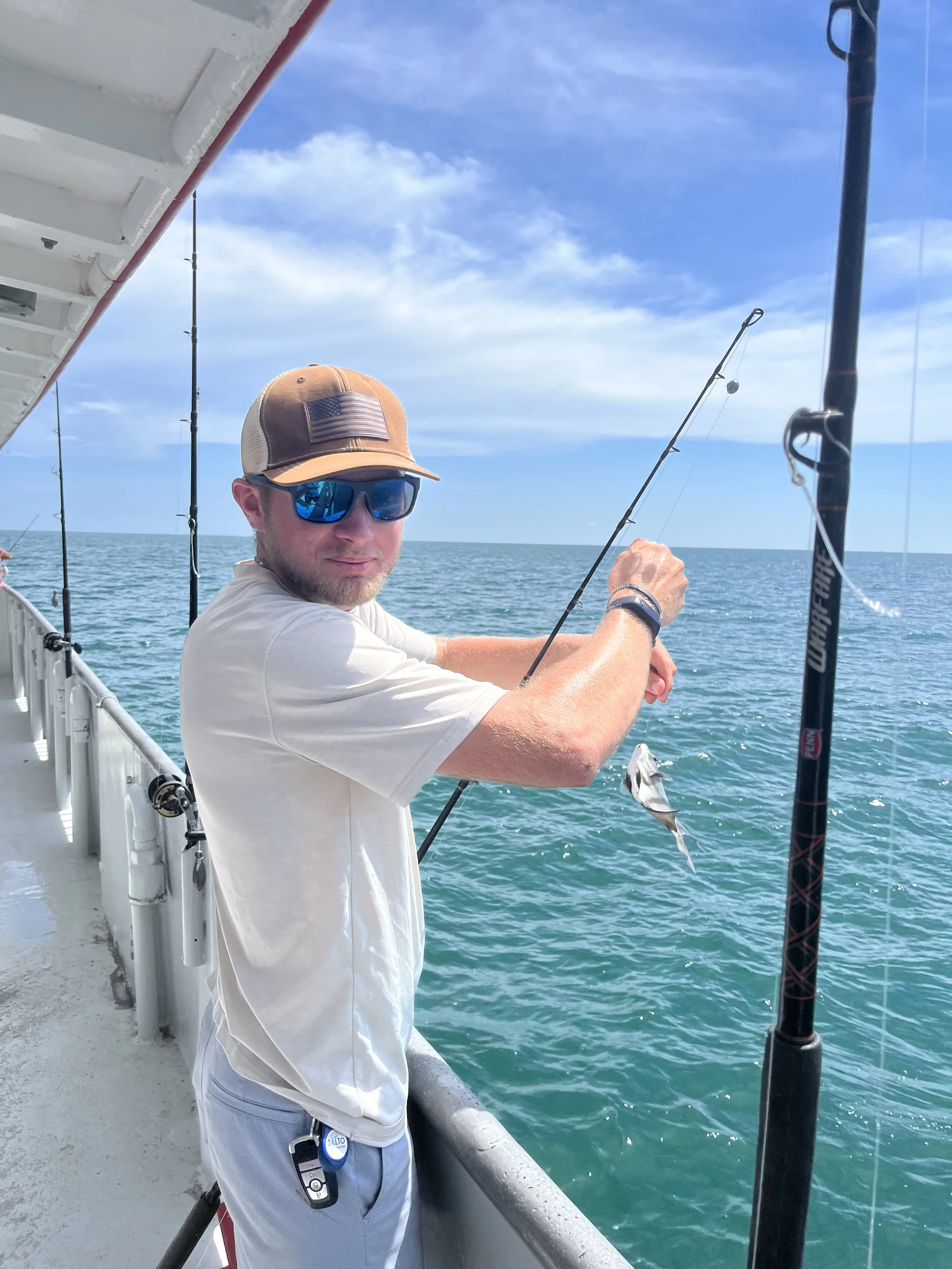 A man with sunglasses and a baseball cap standing on a boat, holding a fishing rod, with the ocean in the background and a blue sky with clouds.