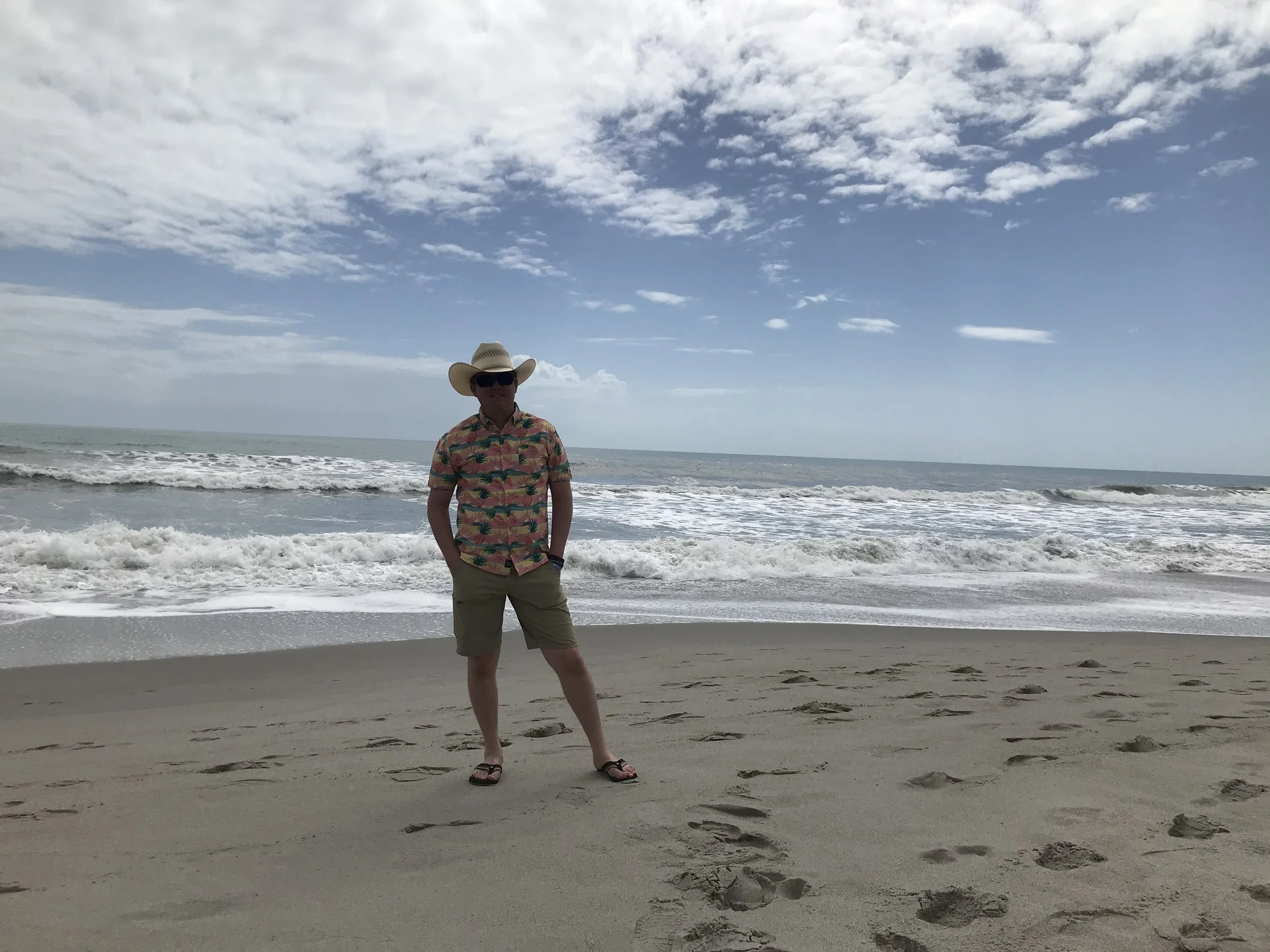 A man standing on the beach near the shoreline with waves, wearing a colorful Hawaiian shirt, shorts, flip-flops, a cowboy hat, and sunglasses, under a sky with scattered clouds.