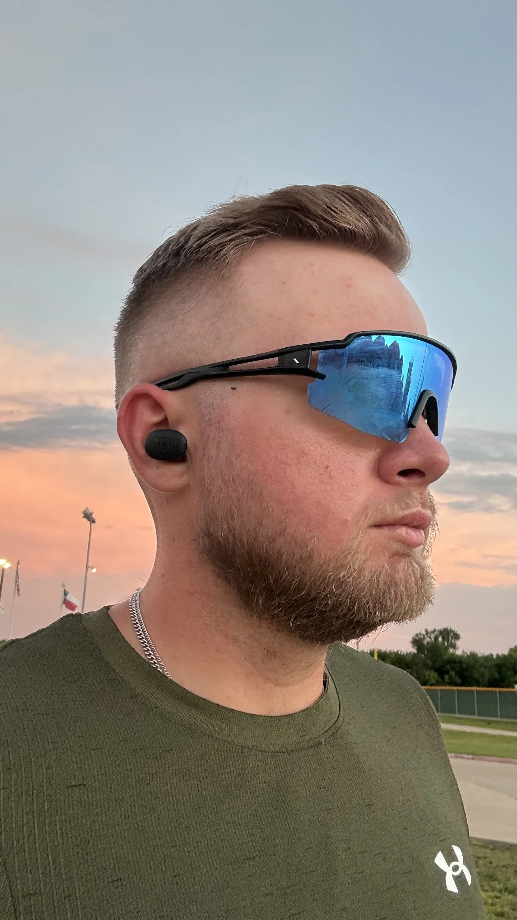 Close-up of a man with sunglasses and earbuds, wearing a green shirt, outdoors during sunset, with flags and trees in the background.