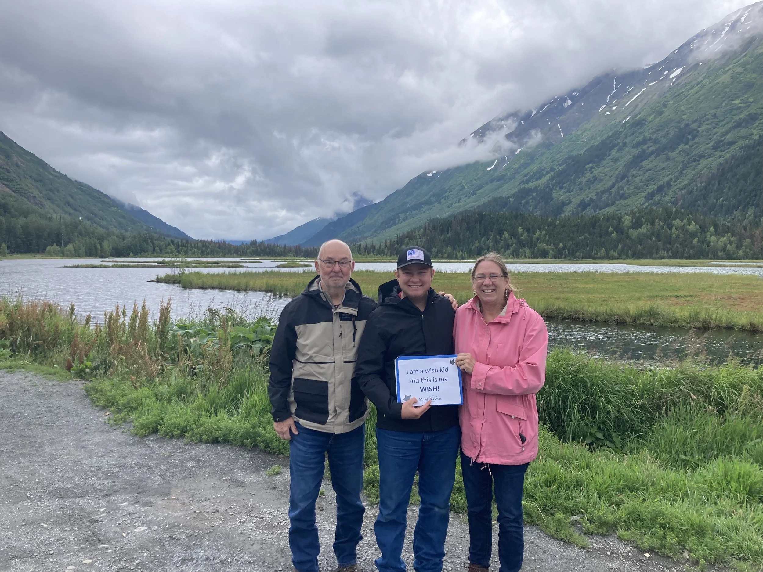 Three people standing outdoors near a lake with green mountains under cloudy sky, holding a sign that reads 'I am a wish kid and this is my WISH!'