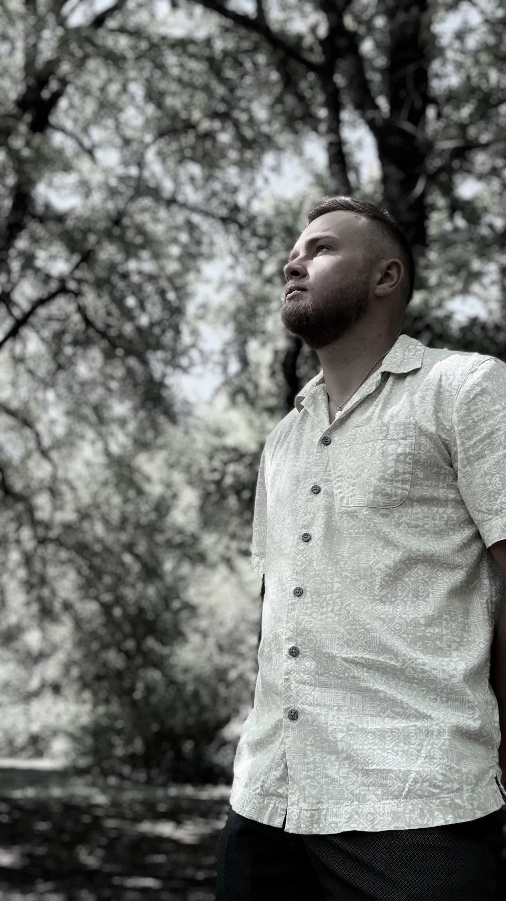 A man with a beard and short hairstyle looking up, standing outdoors in front of trees with leaves and branches, wearing a light-colored patterned short-sleeve shirt.
