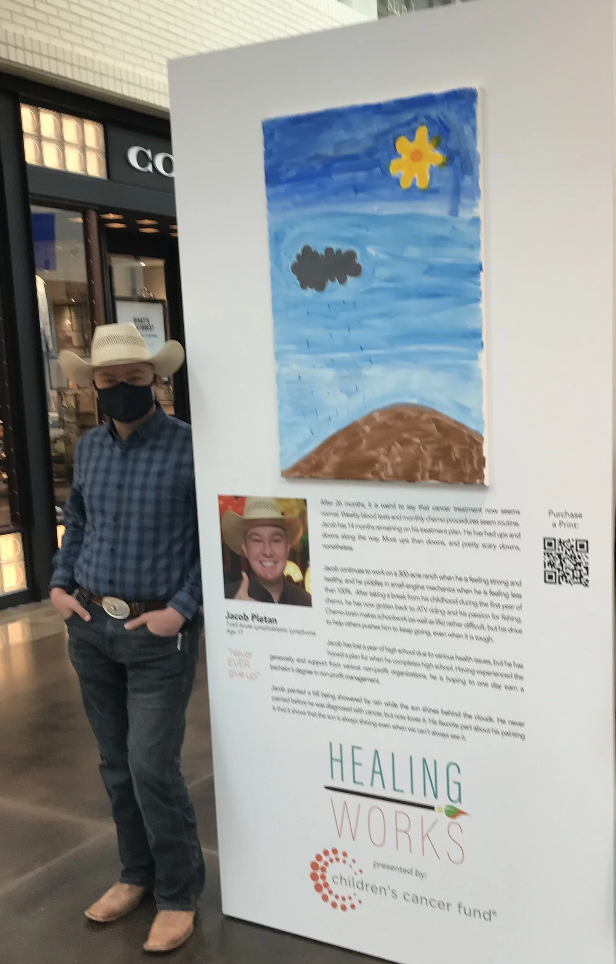 A man wearing a cowboy hat, black face mask, blue plaid shirt, dark jeans, and cowboy boots standing next to an informational poster about healing works supported by Children's Cancer Fund at an indoor venue.