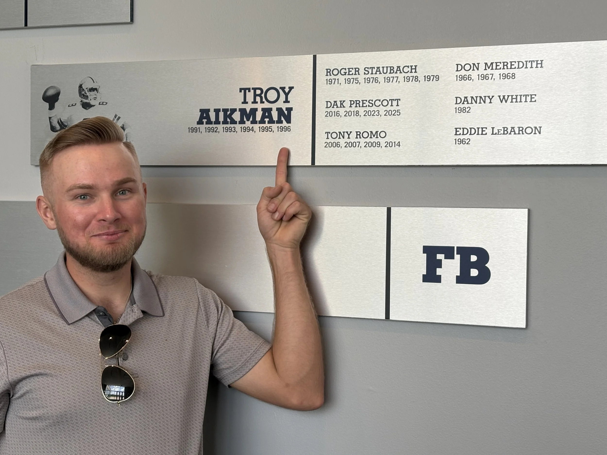 A young man with light brown hair and a beard, wearing a gray polo shirt and sunglasses hanging from his collar, is pointing at a memorial wall. The wall features a black and white image of an American football player in uniform and helmet, along wit