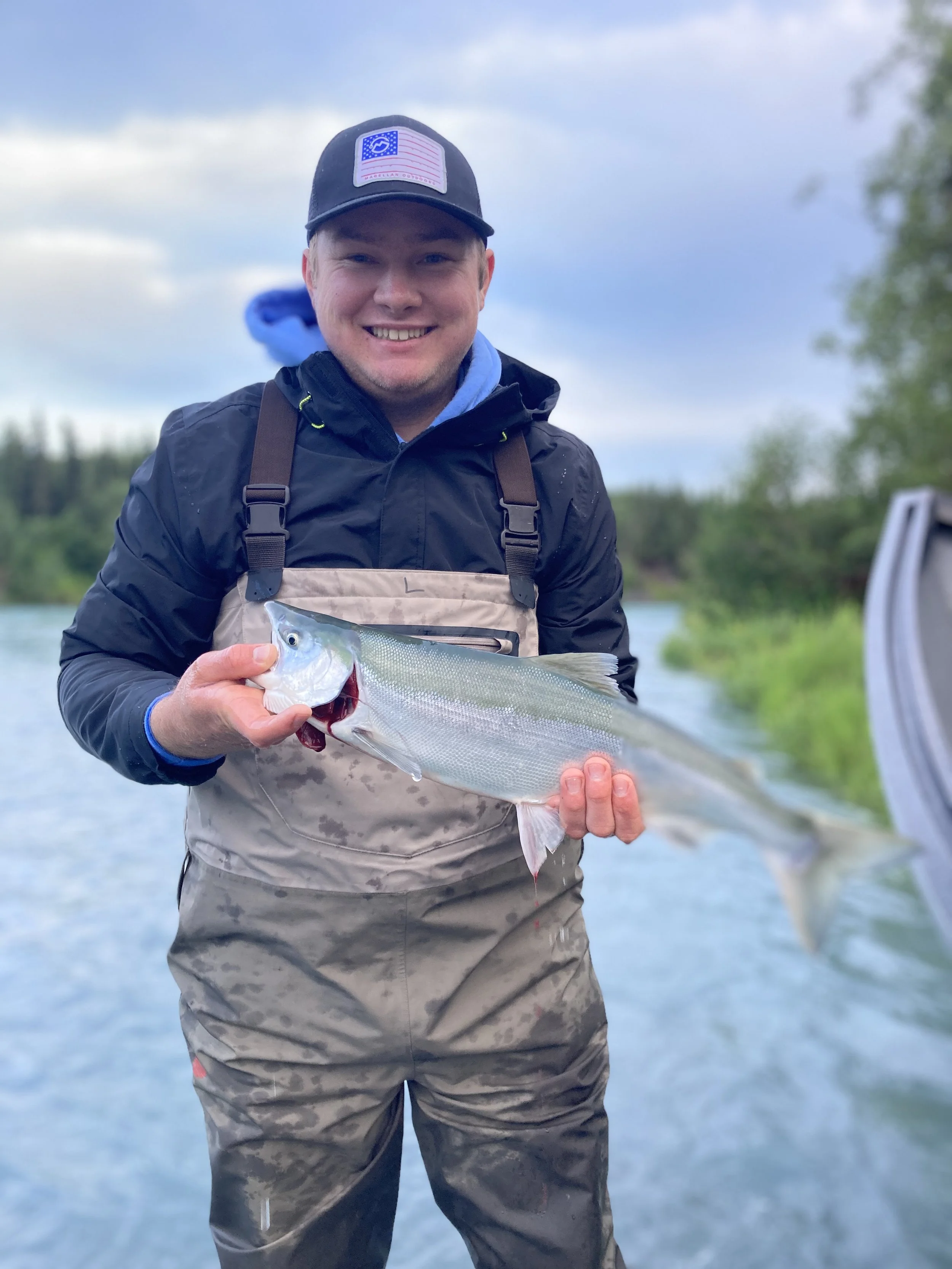 A young man smiling and holding a fish outdoors on a river, wearing waders, a black jacket, and a cap with an American flag patch.