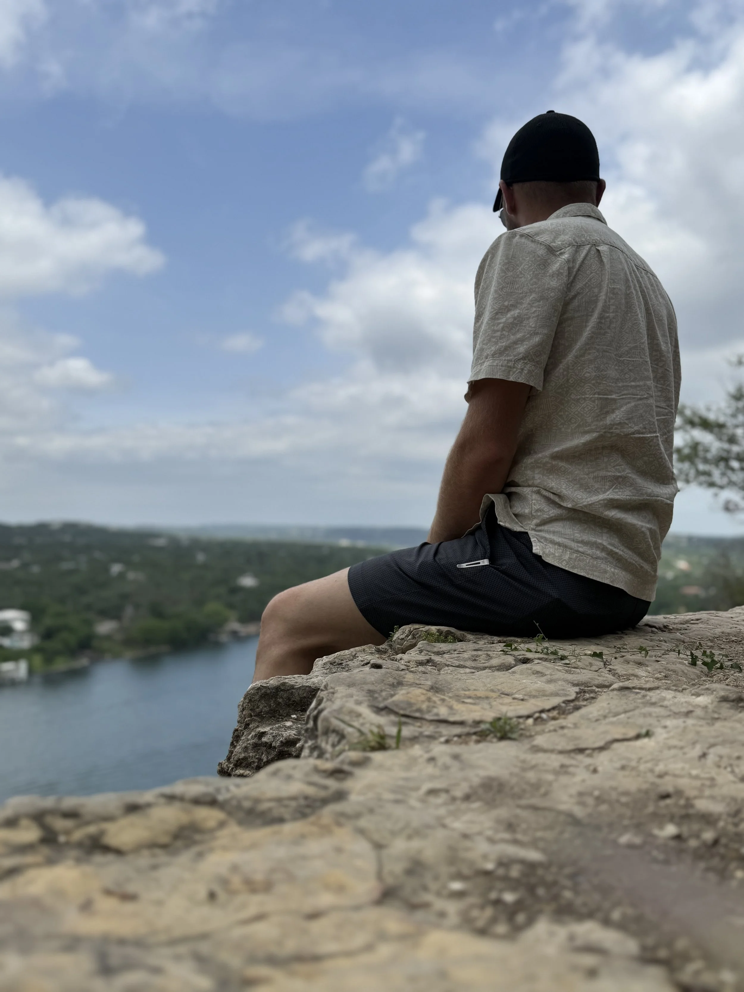 A person sitting on a rocky ledge overlooking a river with trees and buildings in the distance, under a partly cloudy sky.