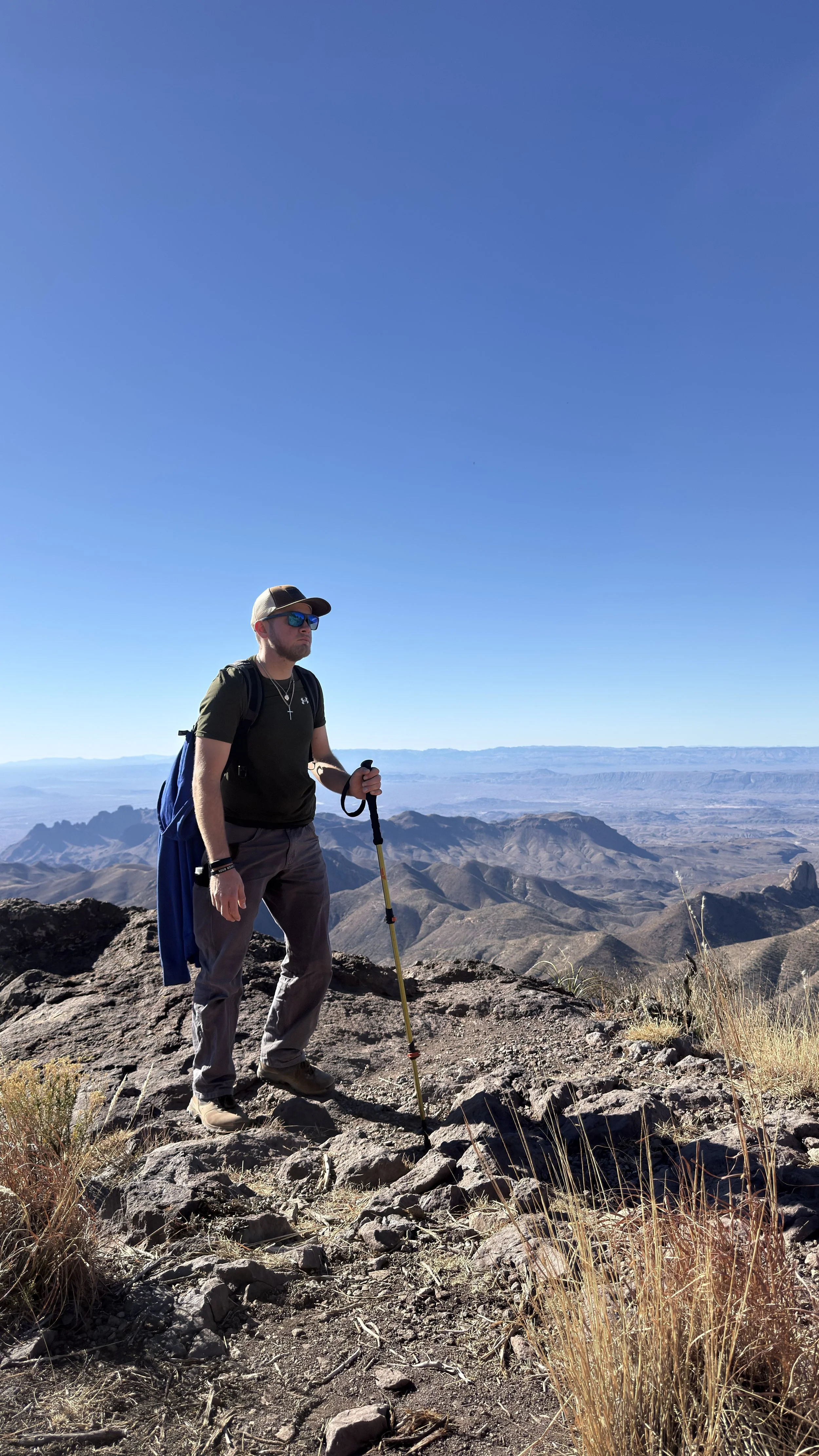 Man standing on rocky mountain trail, holding hiking pole, wearing sunglasses, hat, and backpack, with vast desert landscape and mountains in the background under a clear blue sky.
